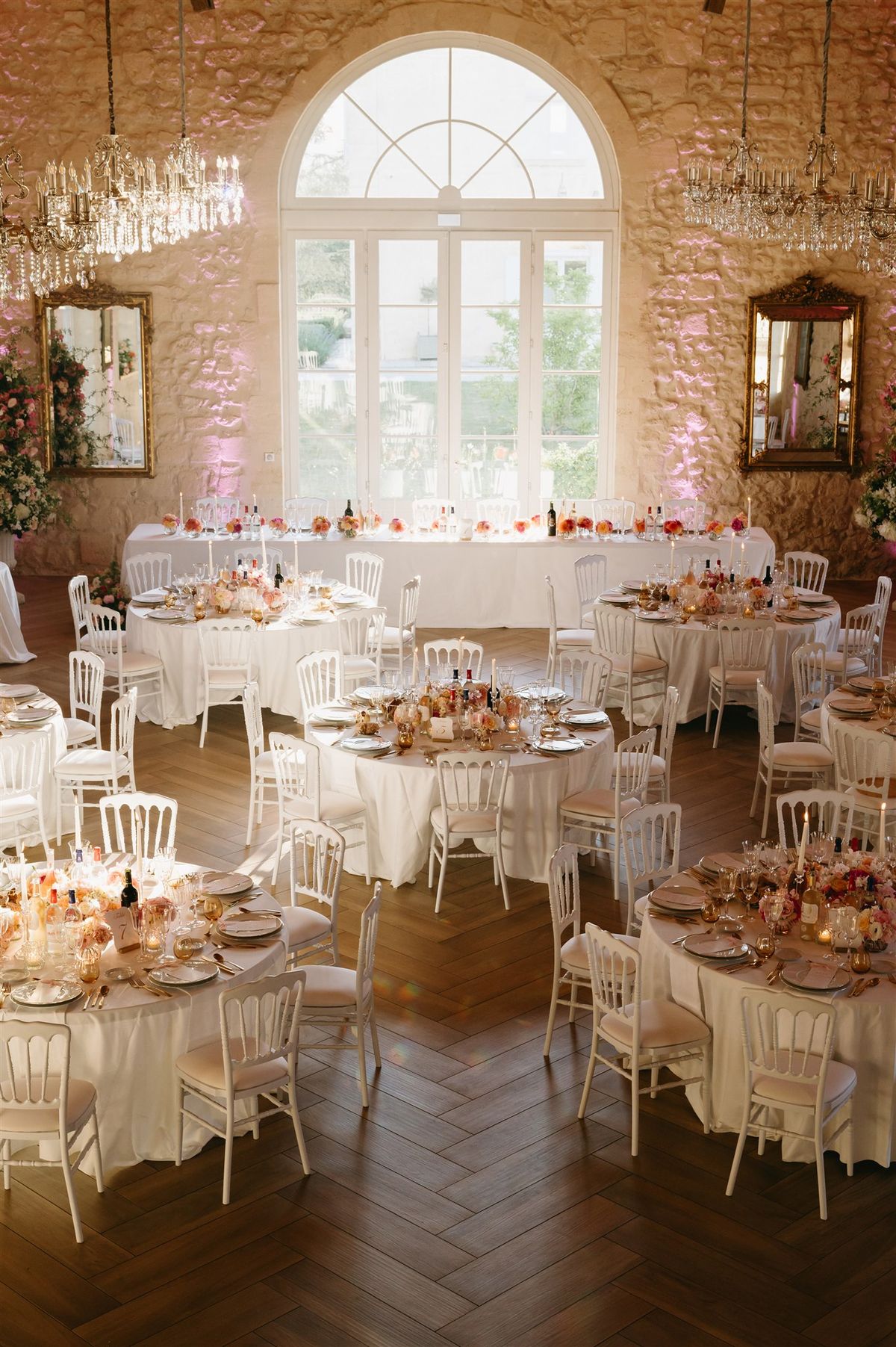 Chateau ballroom with round tables, coral floral centrepieces, Napoleon chairs, and crystal chandeliers