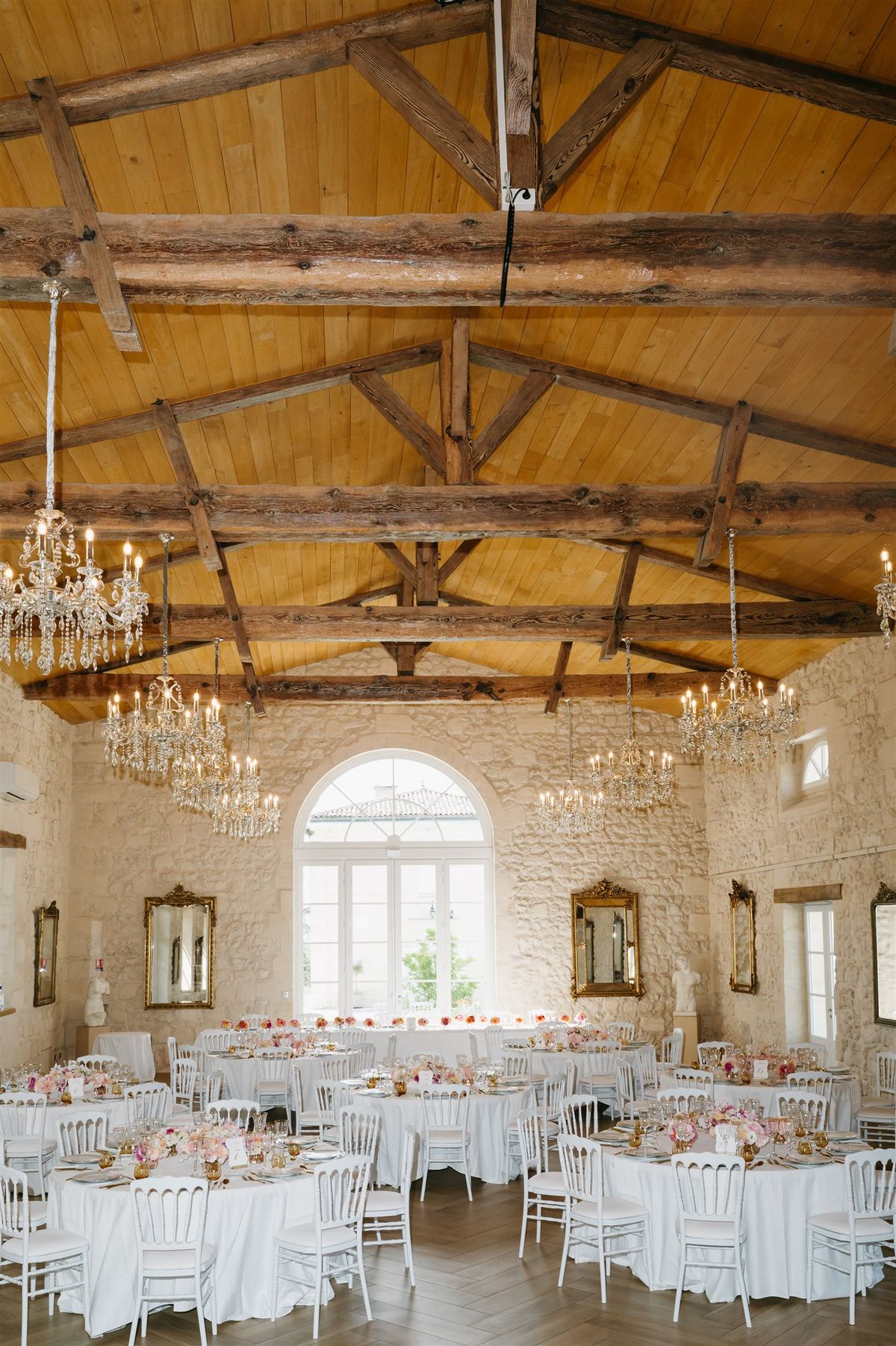 Chateau reception hall with vaulted wood-beam ceiling, crystal chandeliers, round tables with pink and coral floral center...