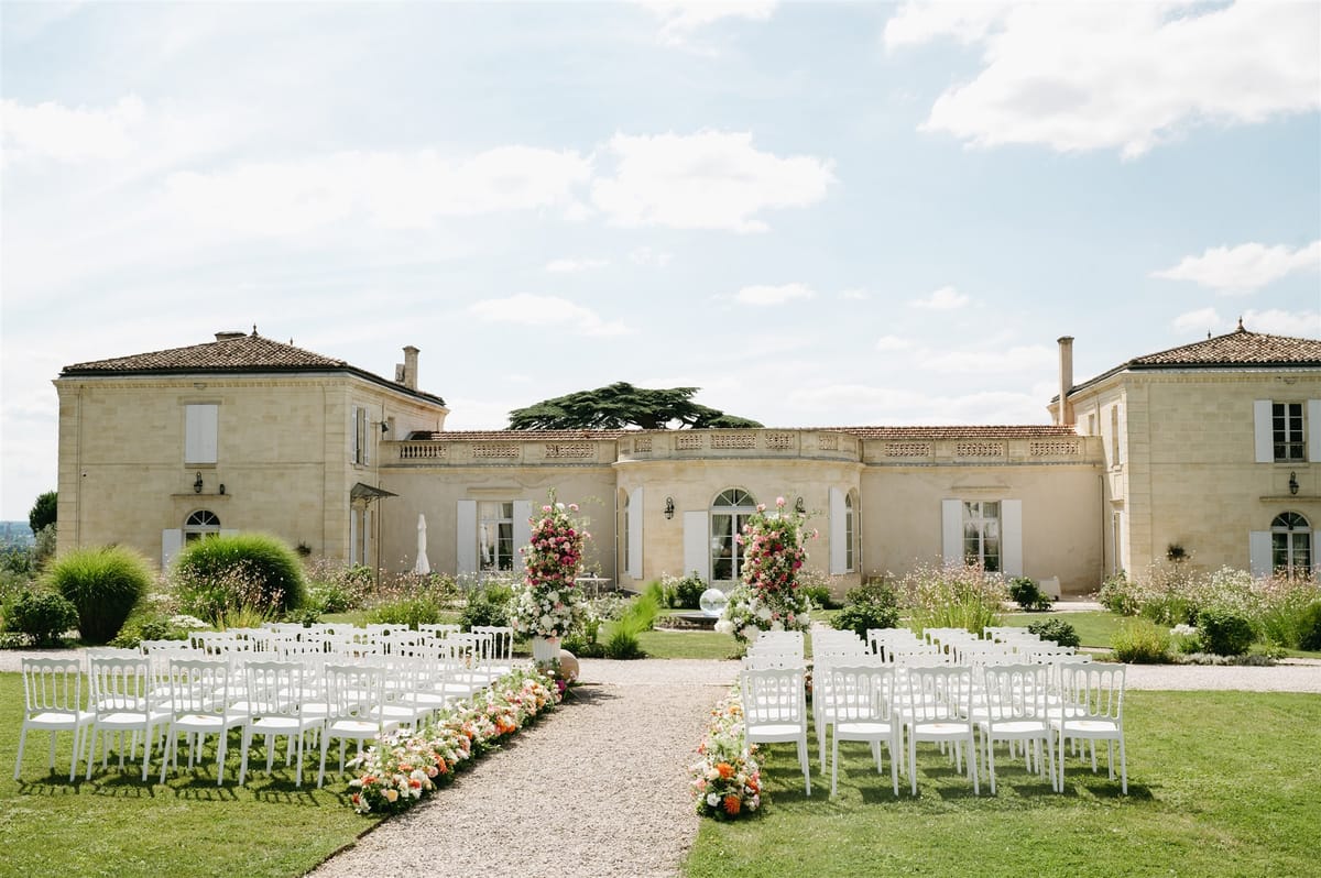 Outdoor wedding ceremony setup with white chairs and coral floral columns in front of a French chateau