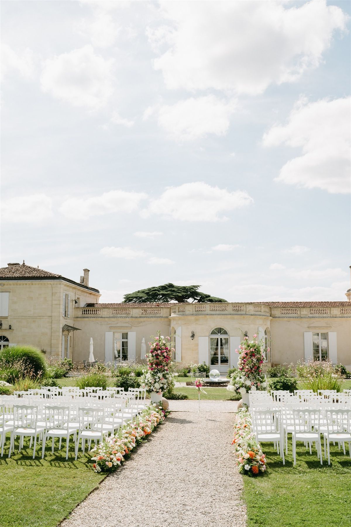 Outdoor ceremony setup with white chairs and colorful floral arrangements on a gravel aisle at a French chateau