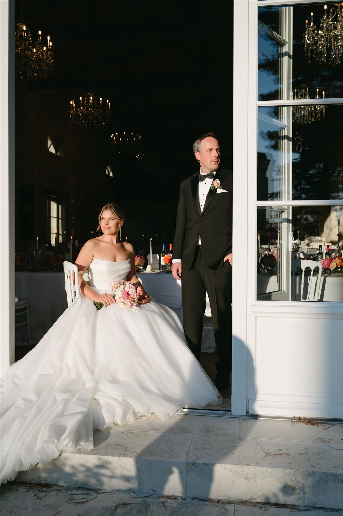 Couple in doorway with bride holding pink and burgundy bouquet, candlelit ballroom with chandeliers behind
