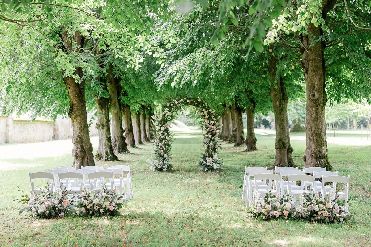Tree-lined ceremony with white chairs, blush rose aisle markers, and circular floral arch