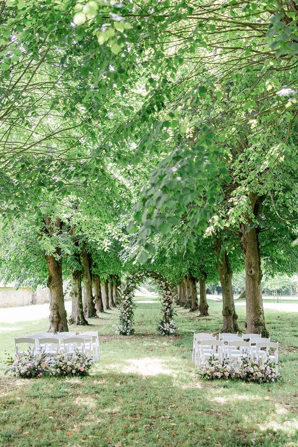 White chairs in tree-lined allee with blush and peach floral installations climbing trunks toward natural canopy