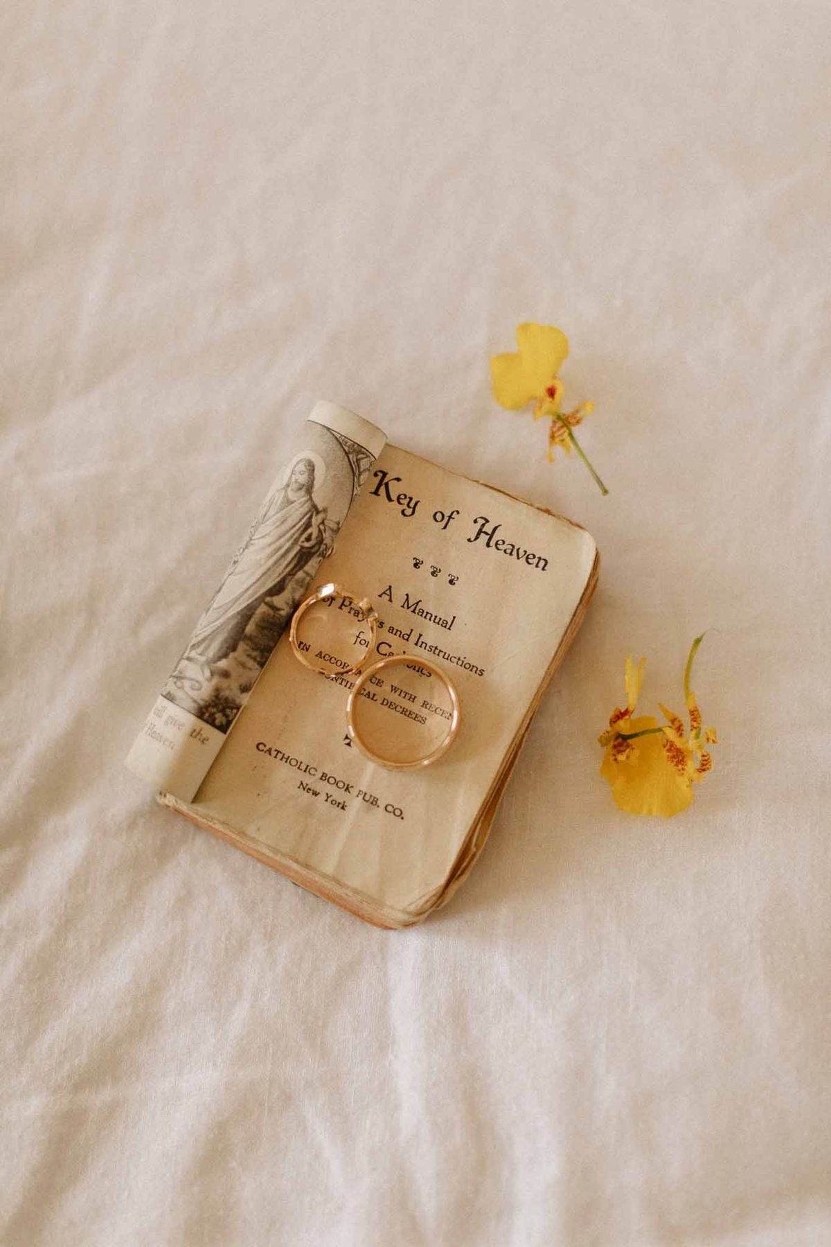 Vintage Catholic prayer book with wedding rings and yellow flowers arranged on cream linen for religious ceremony