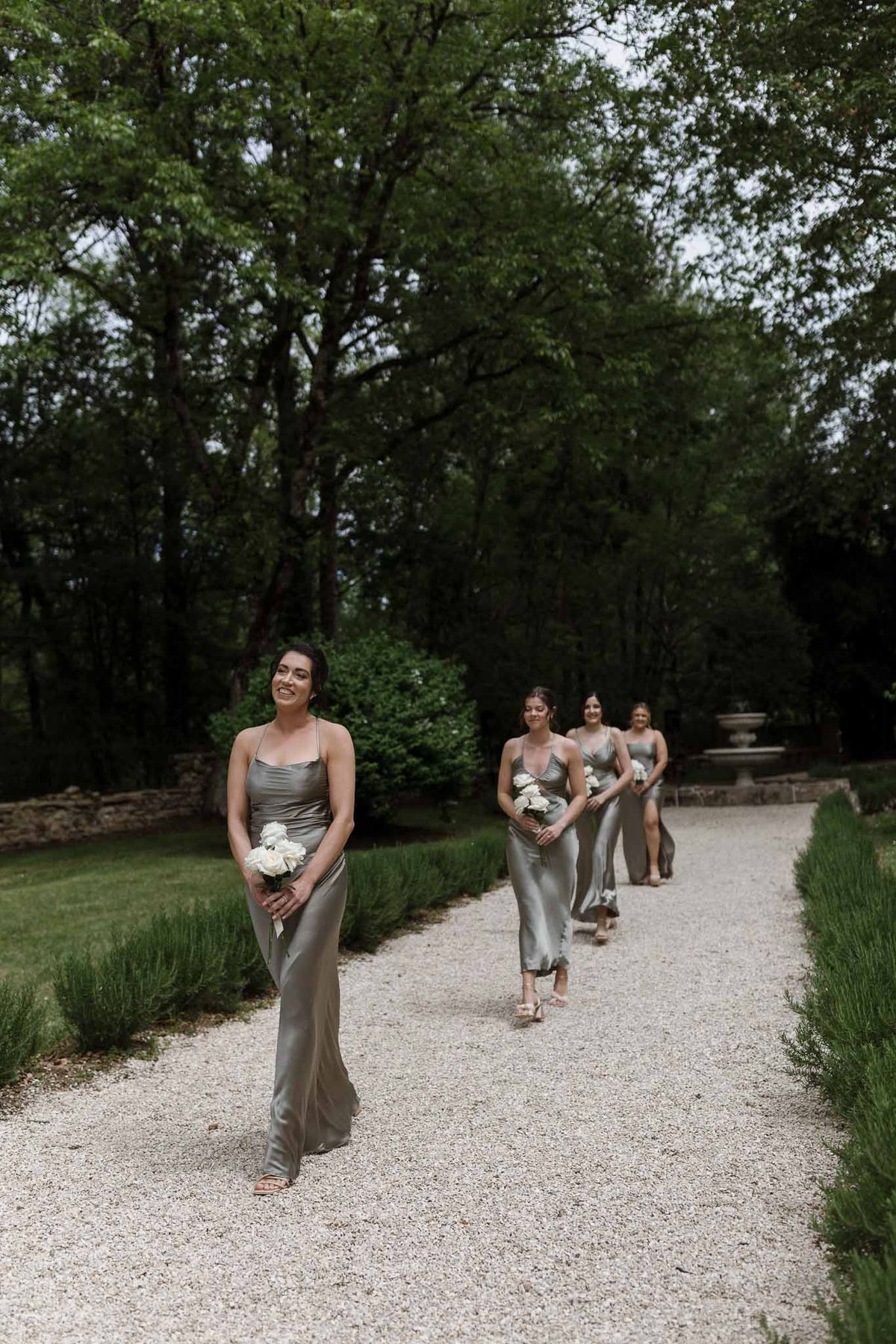 Bridesmaids in sage green dresses walking processional through formal garden with lavender hedging