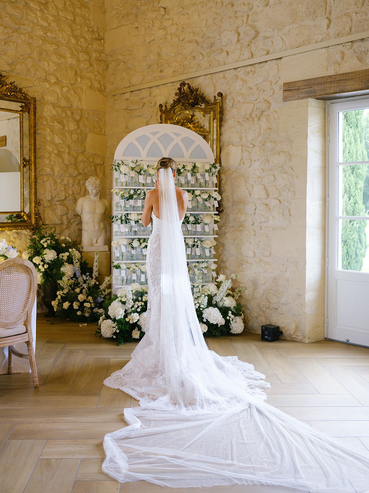 Bride from behind with cathedral lace veil before escort card display with white rose arrangements in limestone room