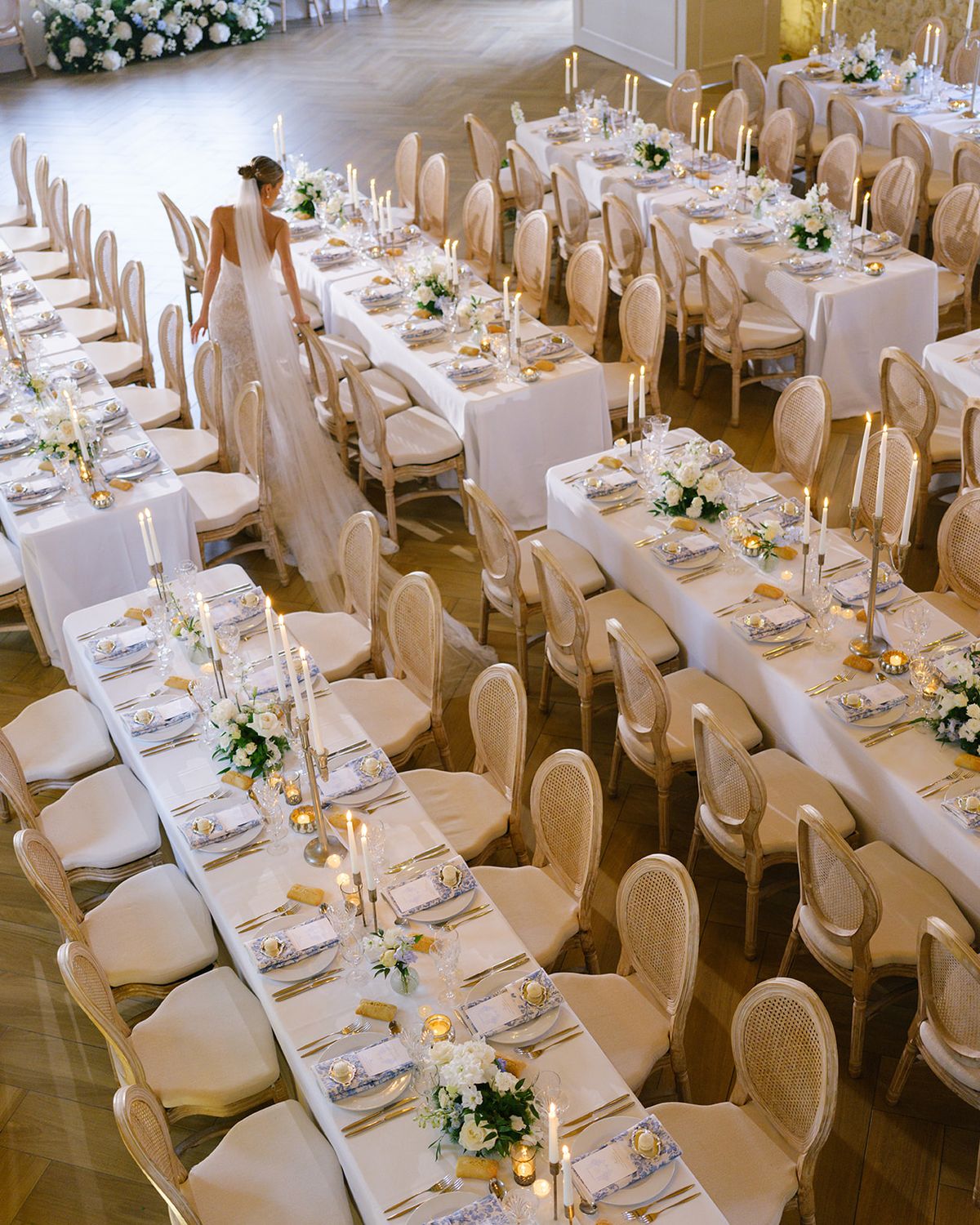 Overhead view of bride walking past banquet tables with gold cutlery, white florals, and cane-back chairs
