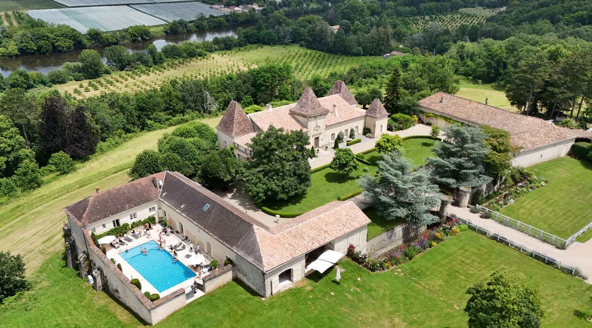 Aerial chateau estate with three conical turrets, pool courtyard, white parasols, and surrounding farmland