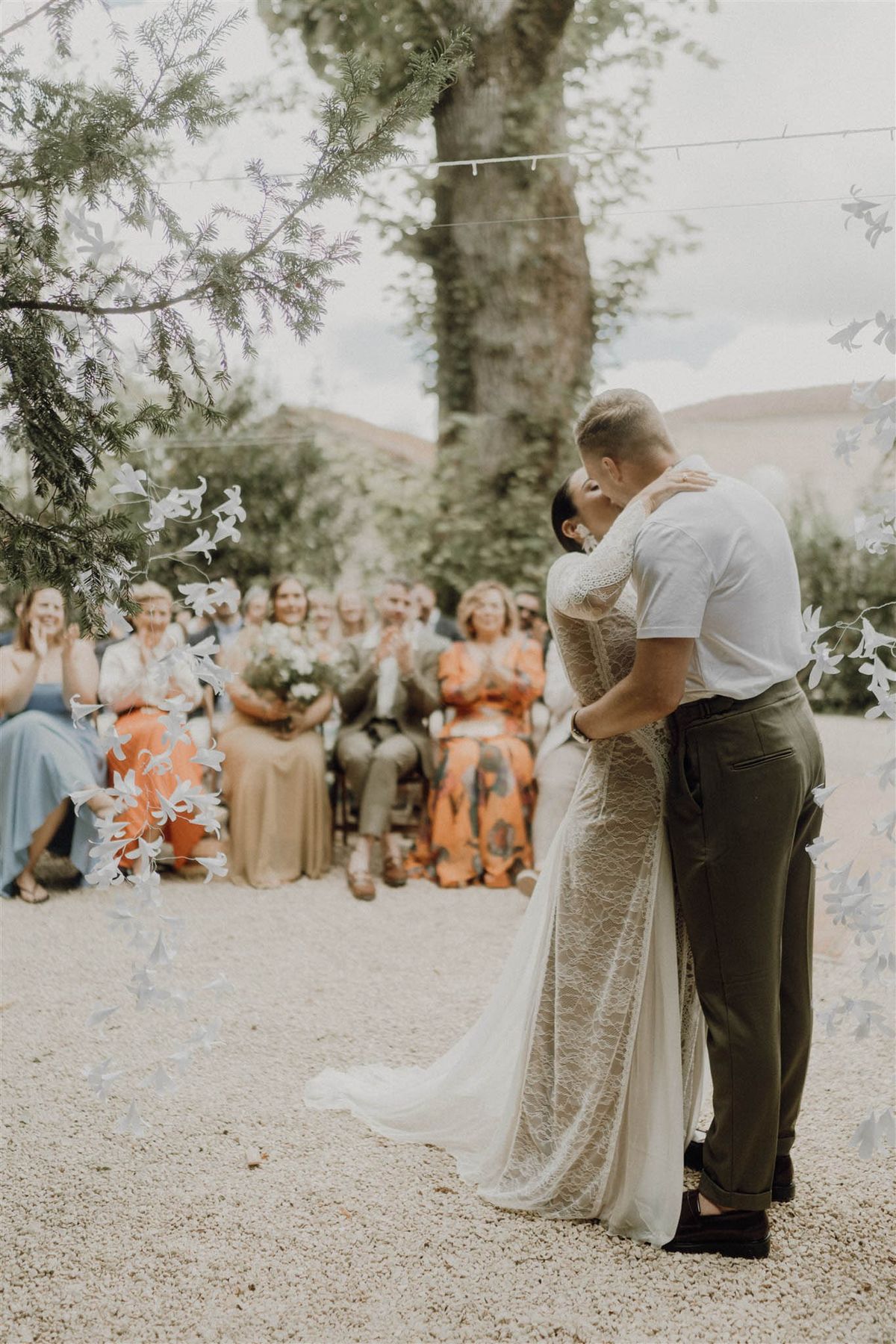 Couple shares first kiss at outdoor ceremony under white paper floral garlands with seated guests applauding behind