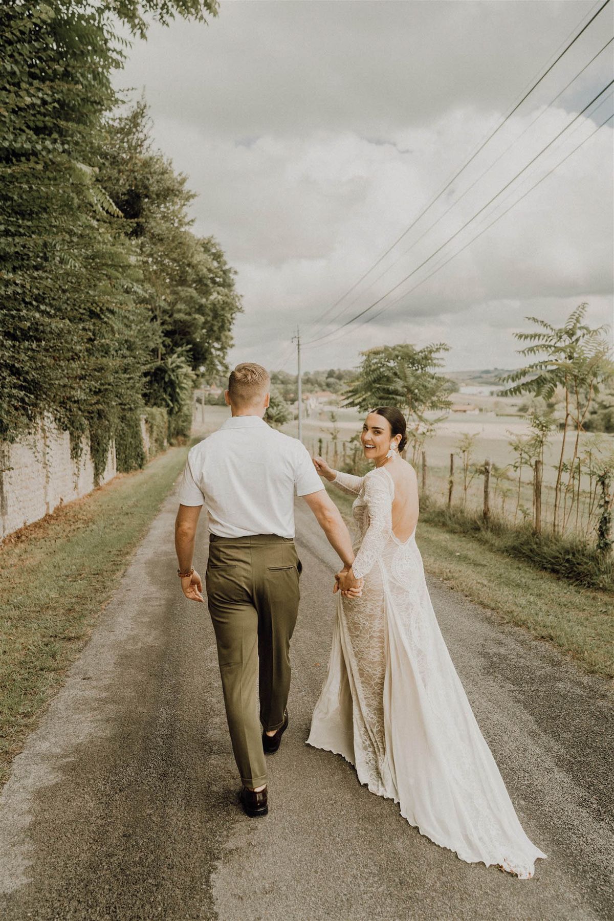 Couple walking away on country road, bride in long-sleeve lace gown turning back with a smile