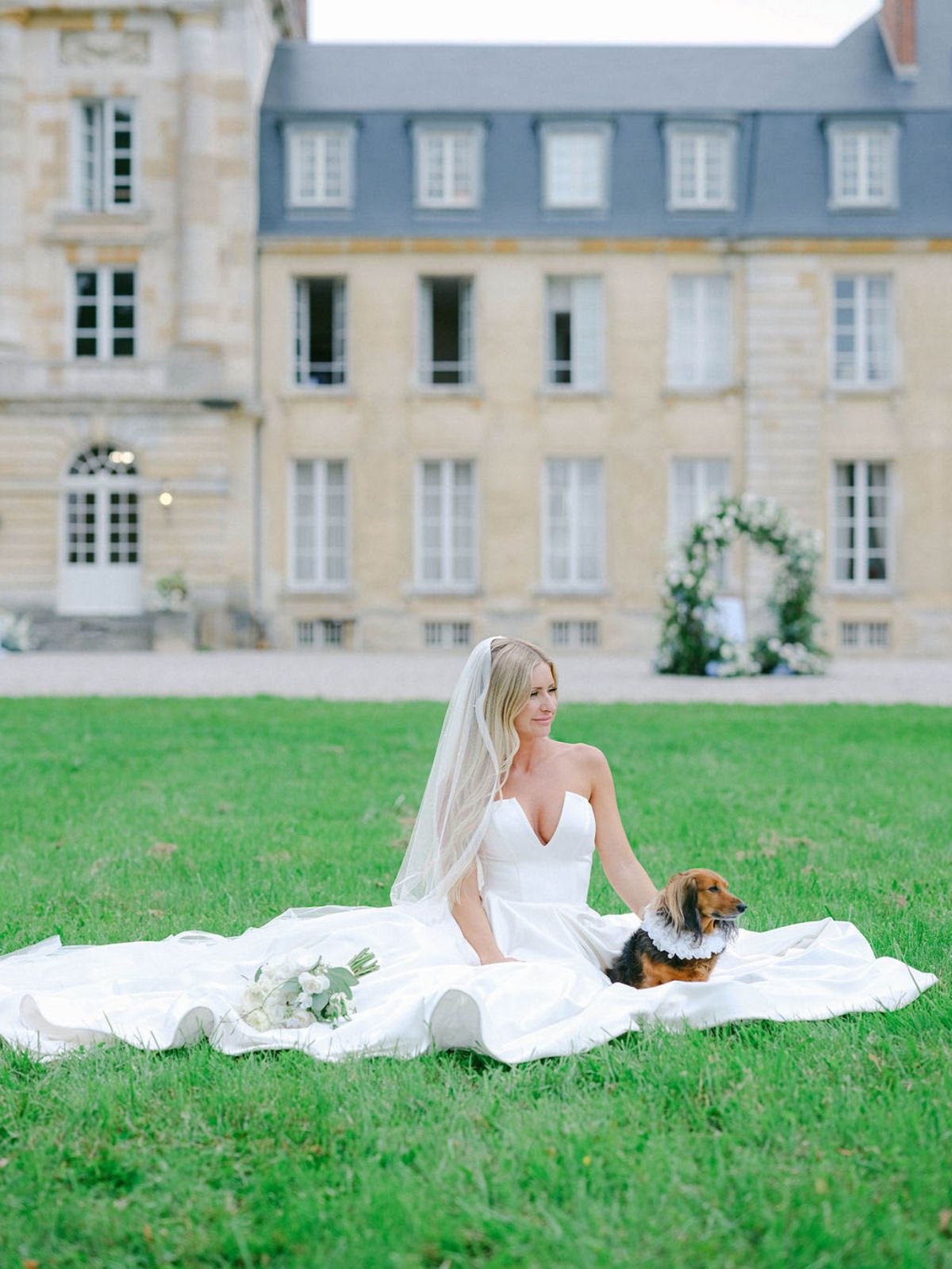 Bride seated on lawn with cathedral veil and dachshund in ruffled collar before honey-stone chateau