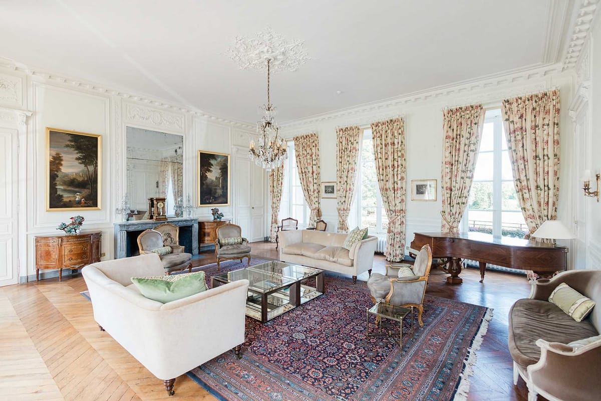 Chateau salon with crystal chandelier, grey marble fireplace, Louis XVI armchairs, and grand piano by windows