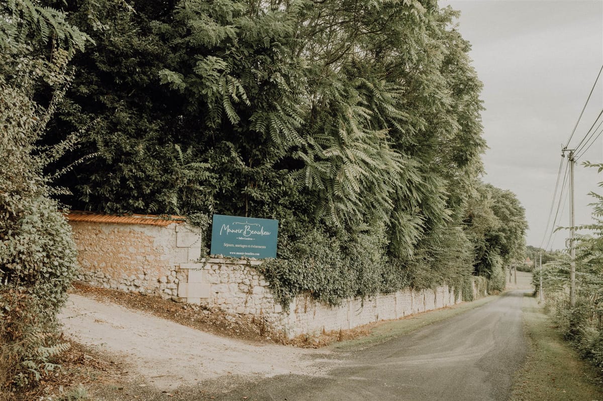 Teal entrance sign for Manoir Beaulieu mounted on limestone boundary wall along a rural road
