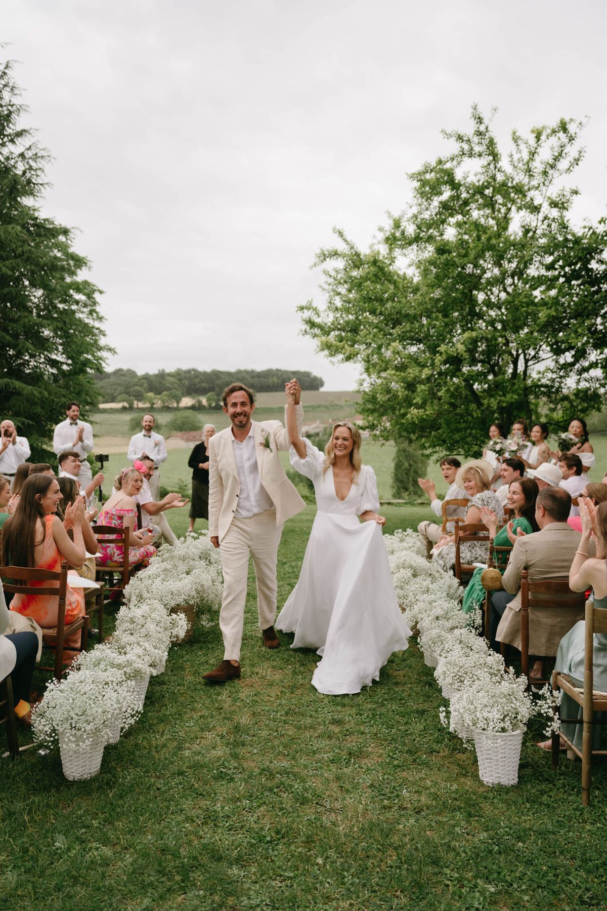 Couple raising hands walking aisle past white gypsophila baskets with cheering guests on lawn