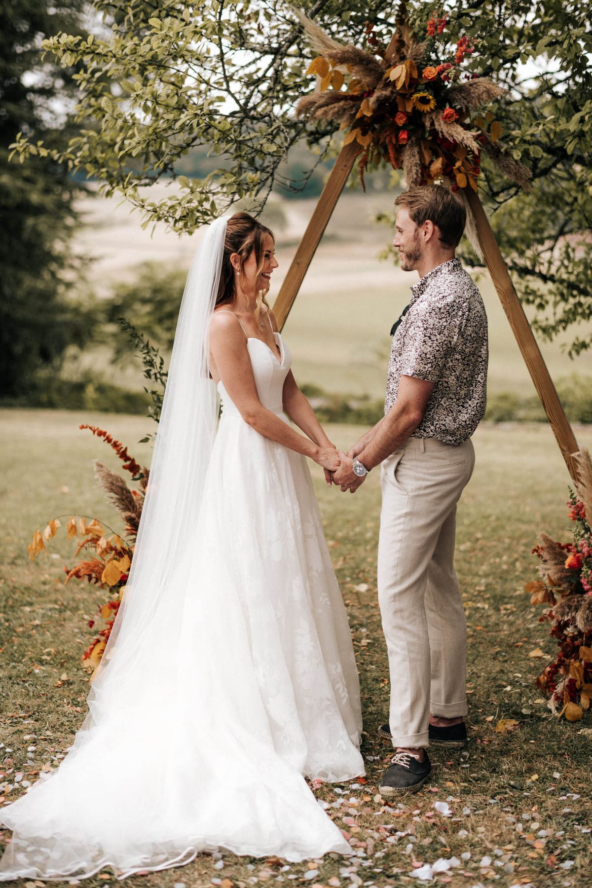 Couple holding hands under triangular wooden arch with pampas grass and sunflower autumn arrangement