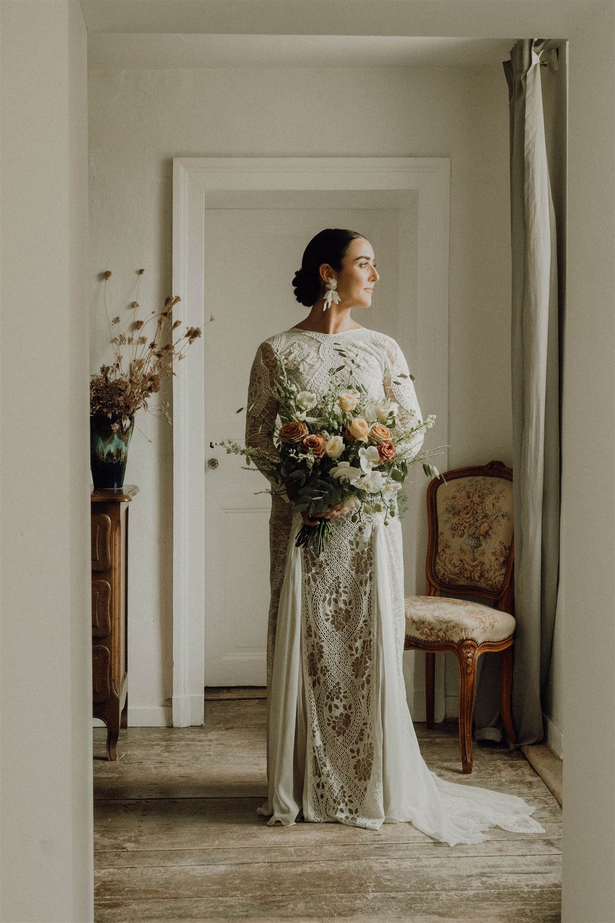 Bride in long-sleeved lace dress holding a garden-style bouquet in a French chateau room with antique furniture
