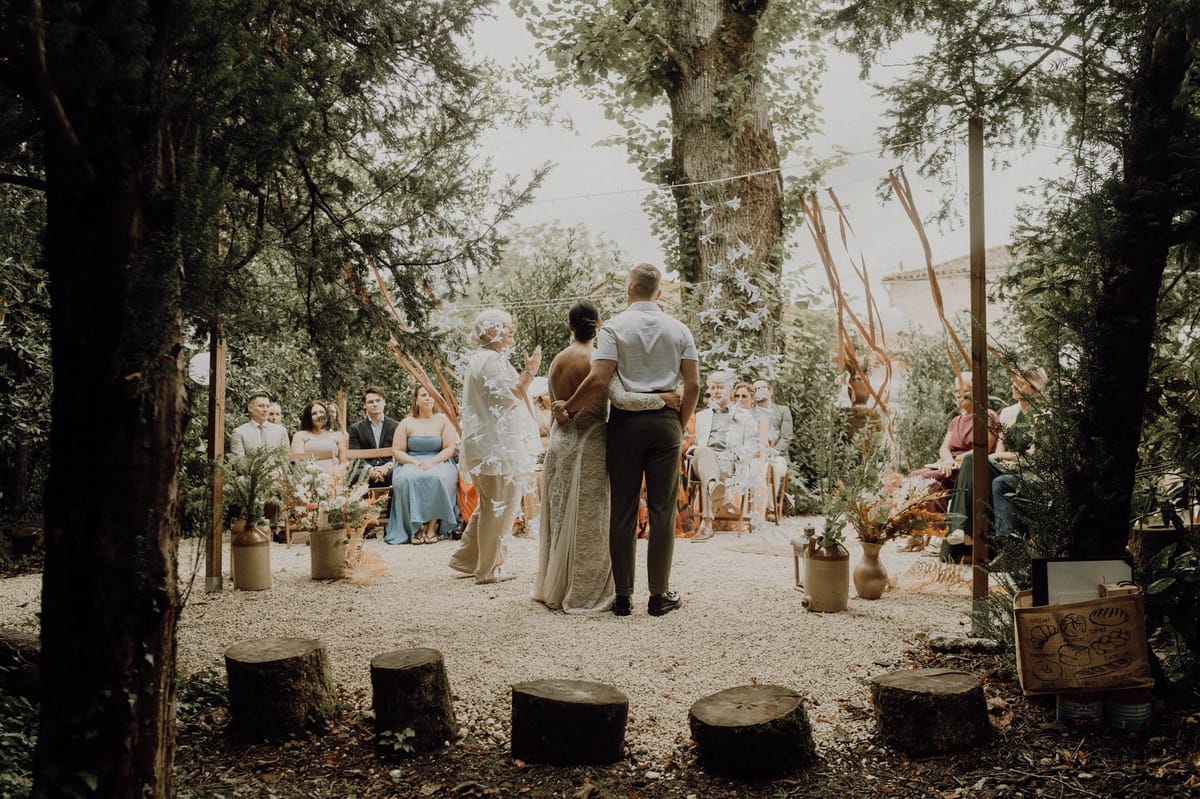 Rustic woodland ceremony with terracotta ribbon streamers, ceramic jug florals, and 20 guests on wooden chairs