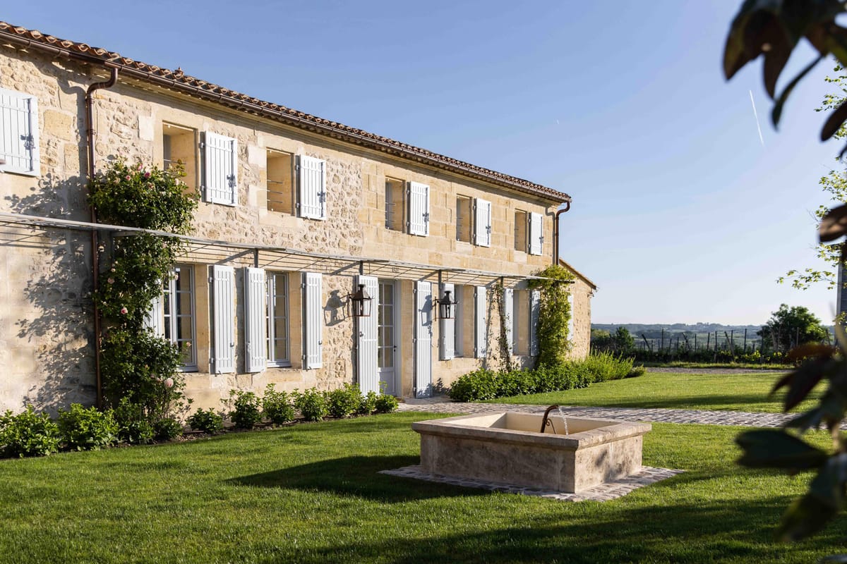 French stone manor house with white shutters, metal pergola with iron lanterns, and stone water trough on lawn