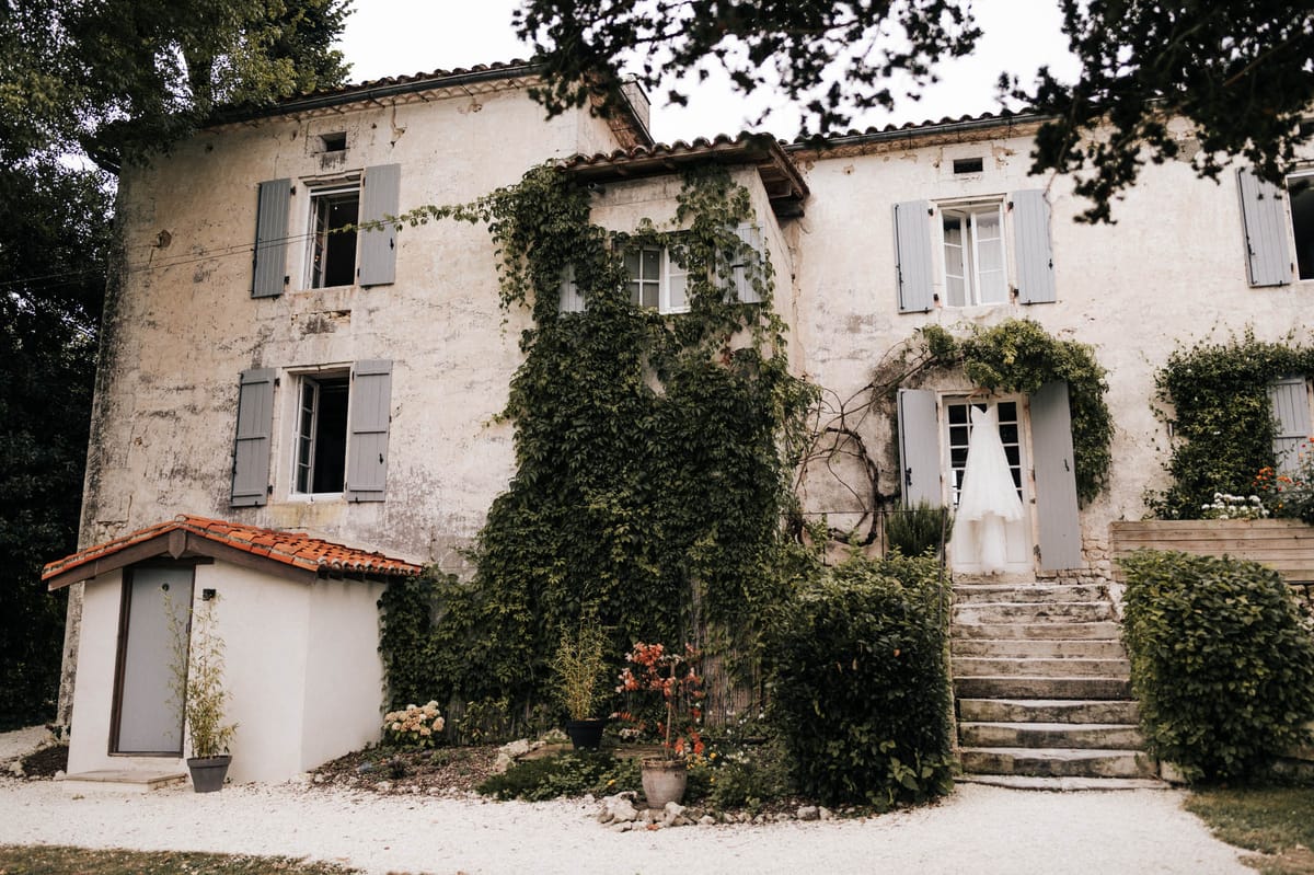 Ivy-covered white manor with blue shutters and wedding dress hung in doorway on gravel forecourt
