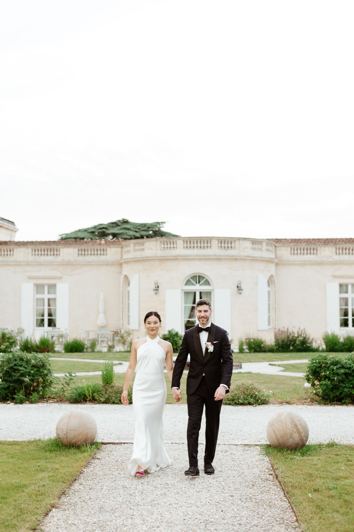 Couple walking hand in hand on gravel path before cream limestone chateau with formal garden