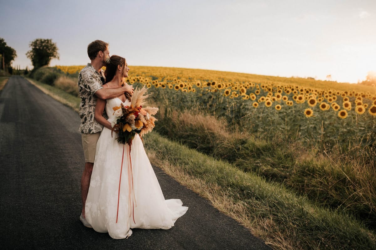 Groom embracing bride from behind on rural road with sunflower field at golden hour in Dordogne