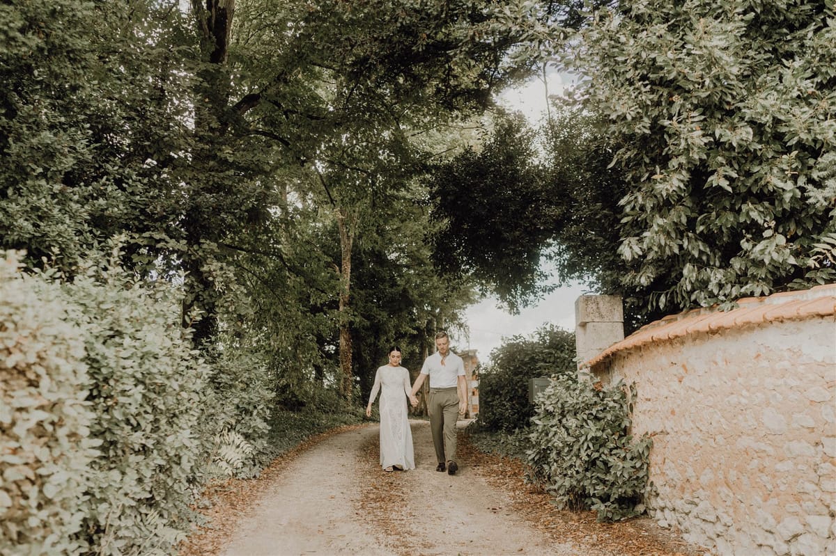 Bride in ivory lace gown and groom walking hand in hand along a hedge-lined gravel path under tree canopy