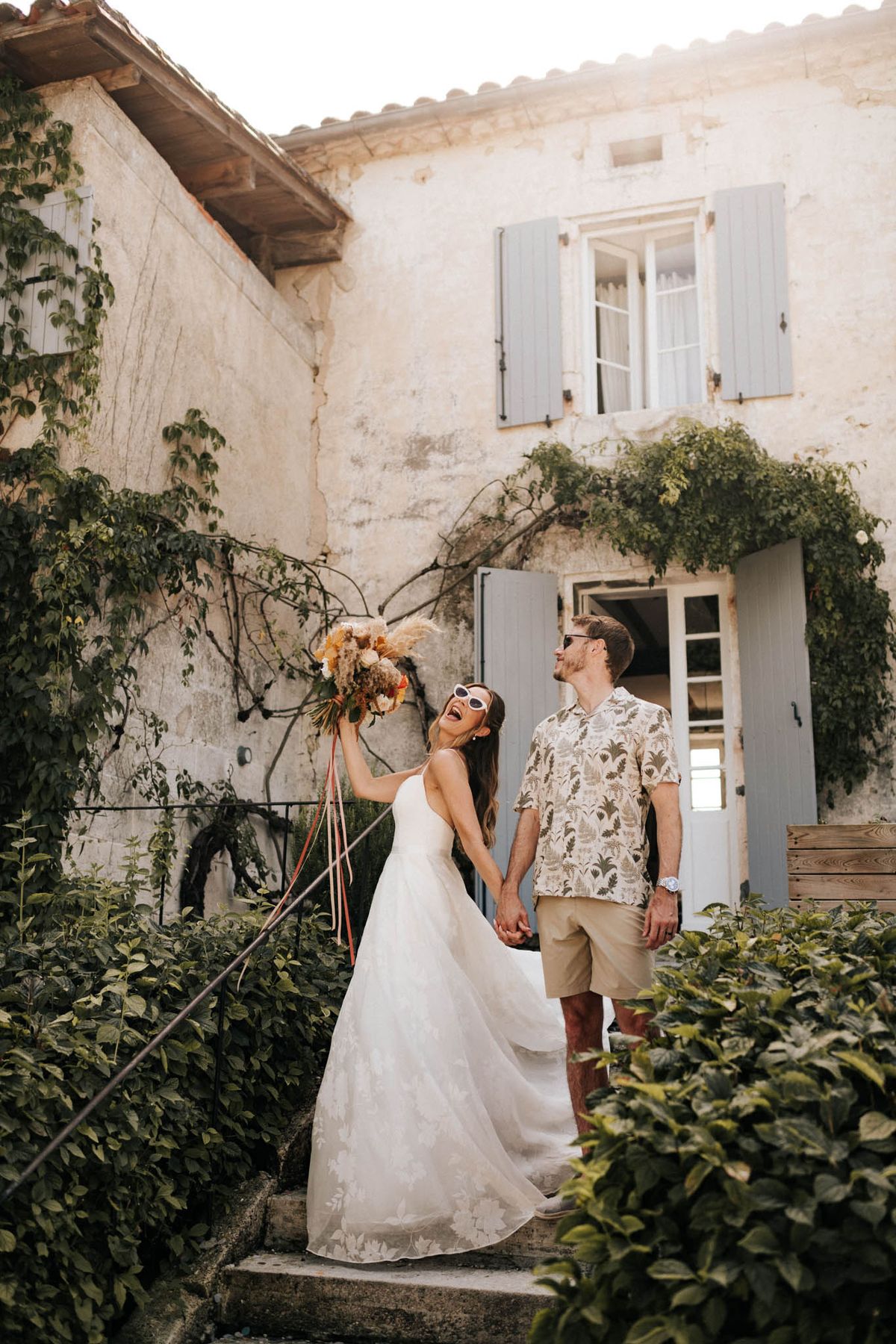 Bride in white A-line gown and groom in tropical shirt laughing on stone steps of French farmhouse with blue shutters