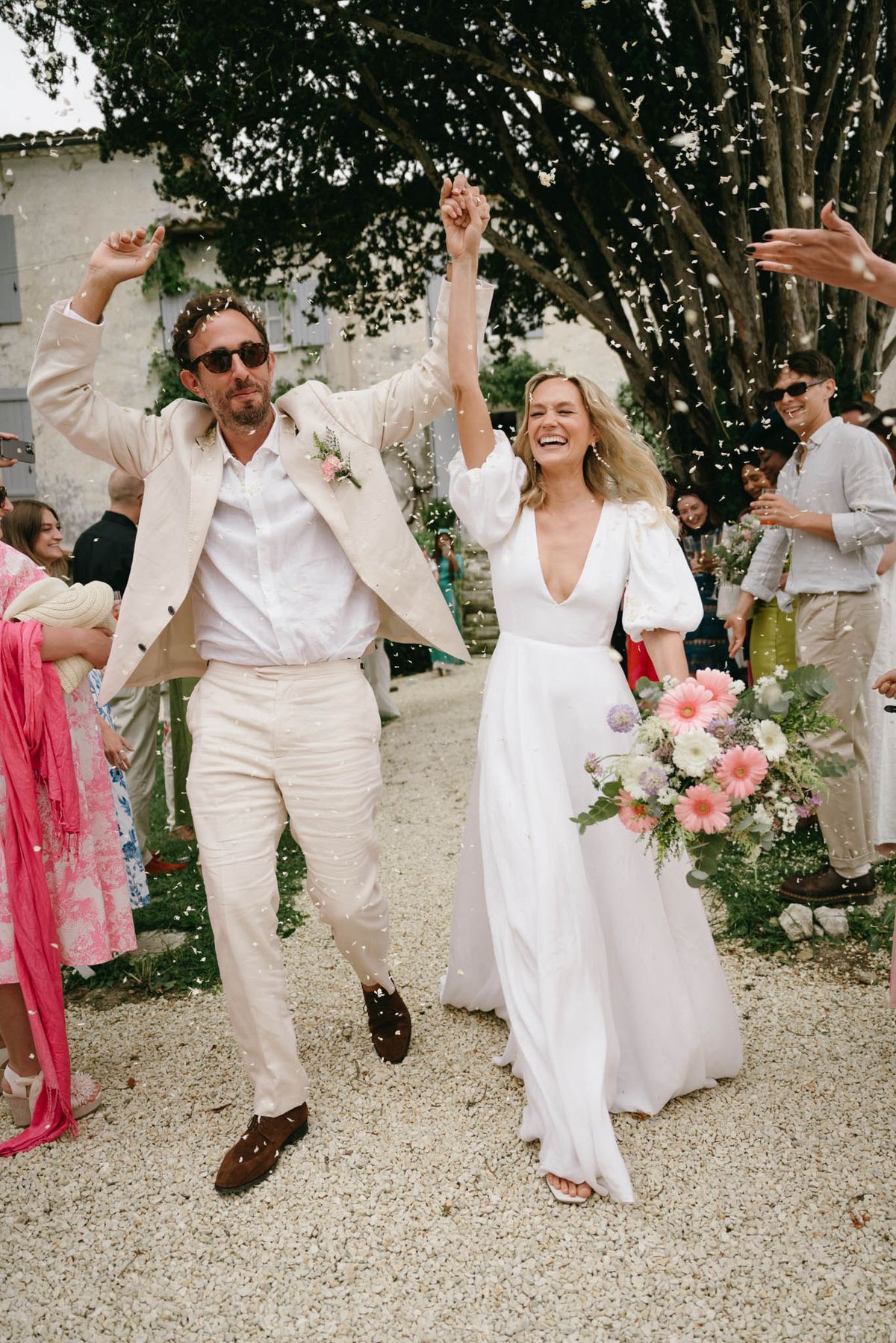 Couple mid-jump through white confetti exit, bride with pink gerbera bouquet, groom in cream linen suit