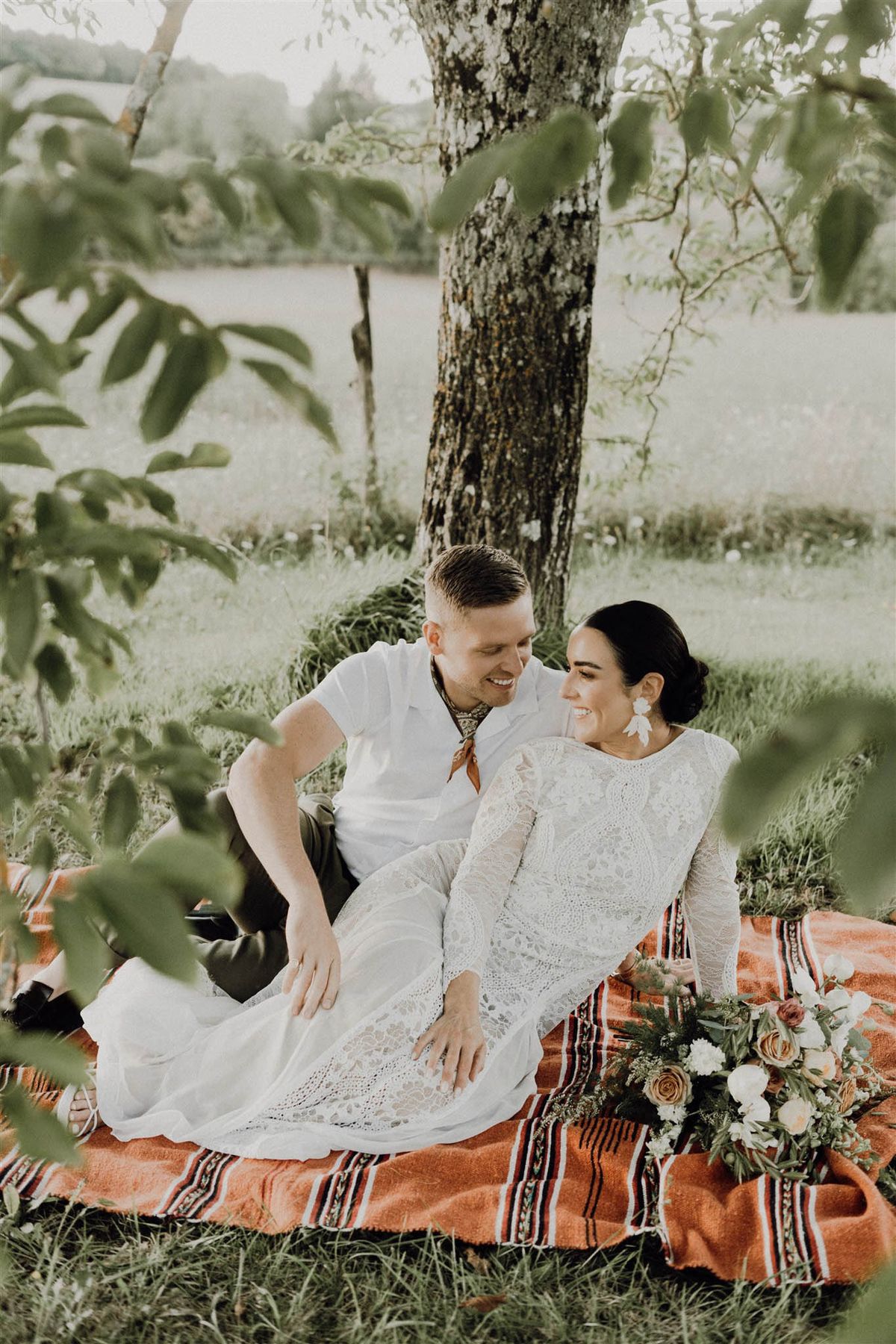 Bride in ivory lace boho gown and groom in white shirt seated on striped blanket under a tree smiling at each other