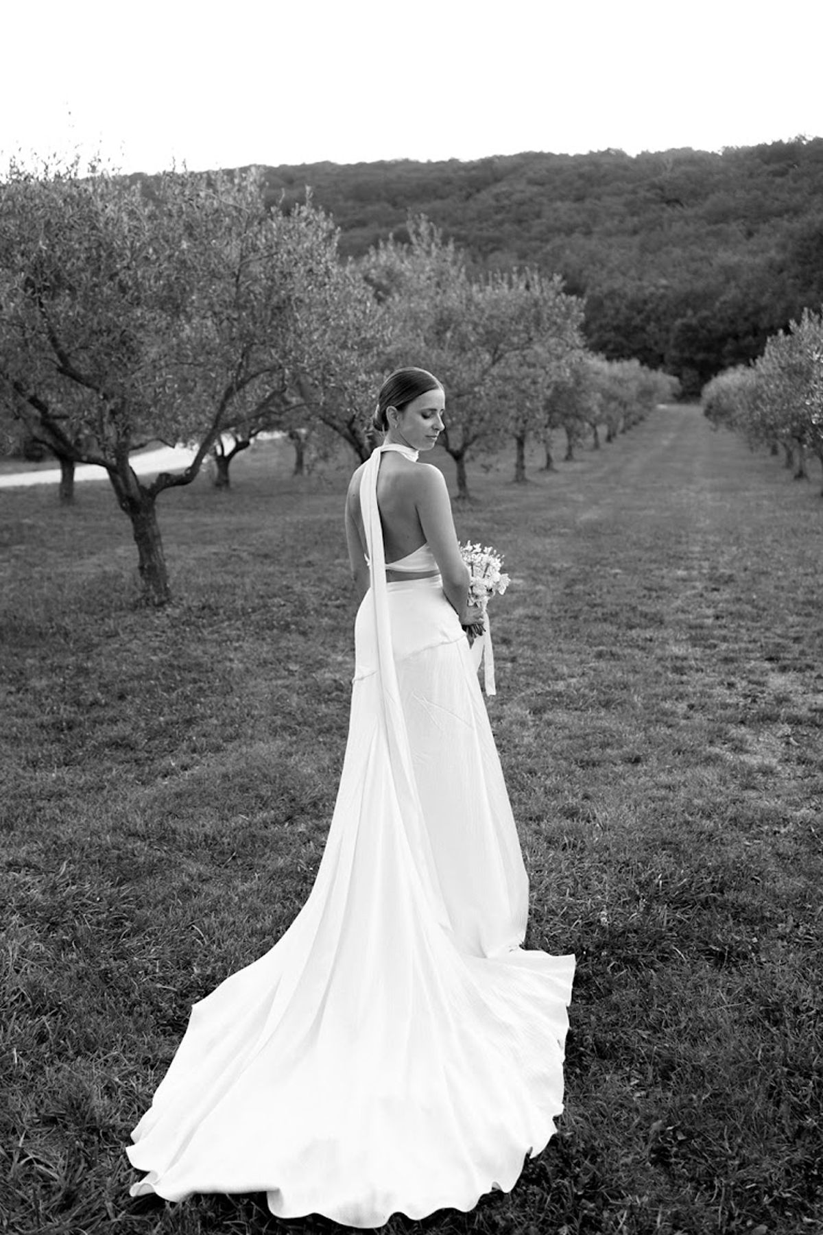 Black and white portrait of bride from behind in open-back halter gown walking through an olive grove