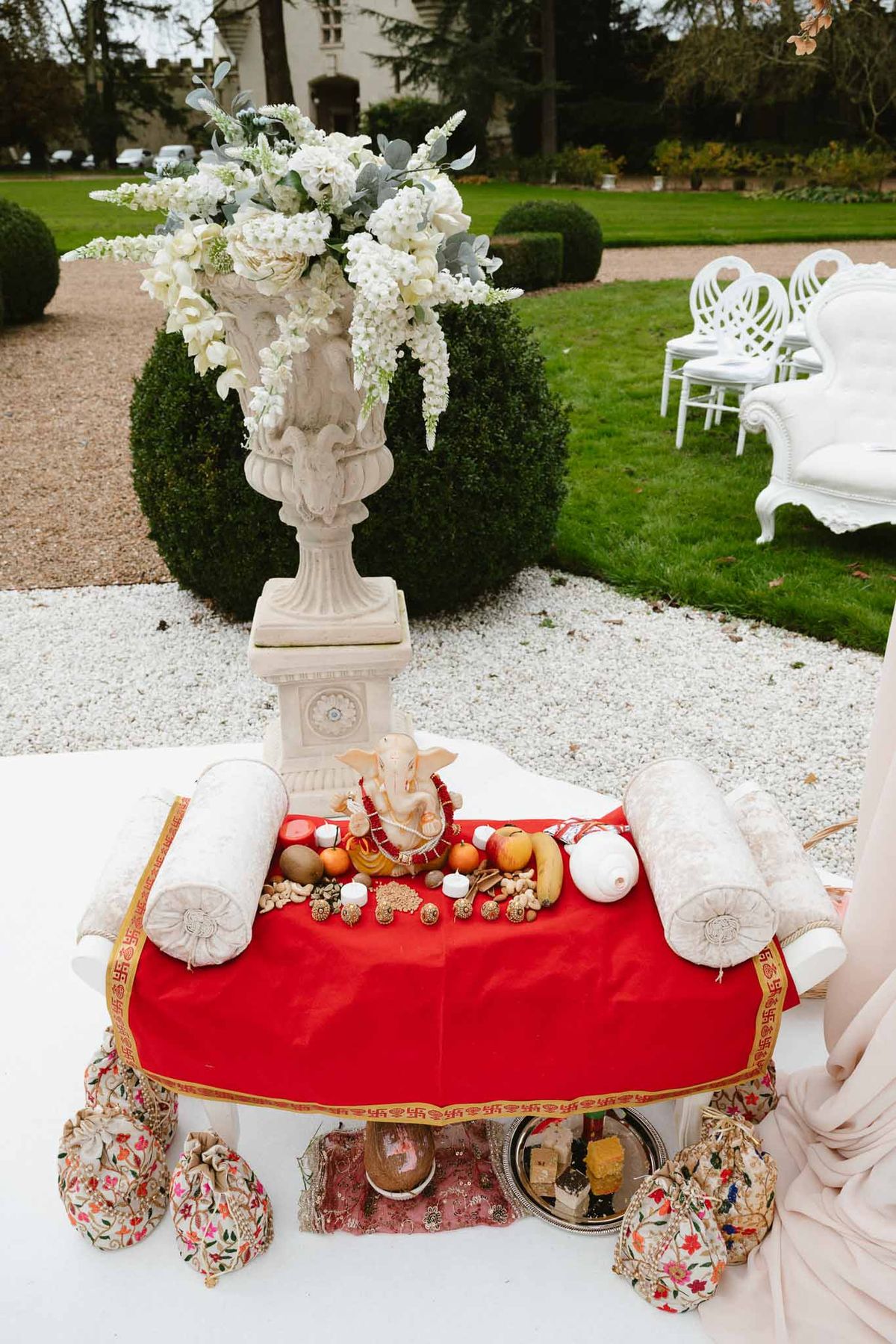 Multicultural wedding ceremony altar table with red fabric, ivory flowers, and gold accents in formal garden setting