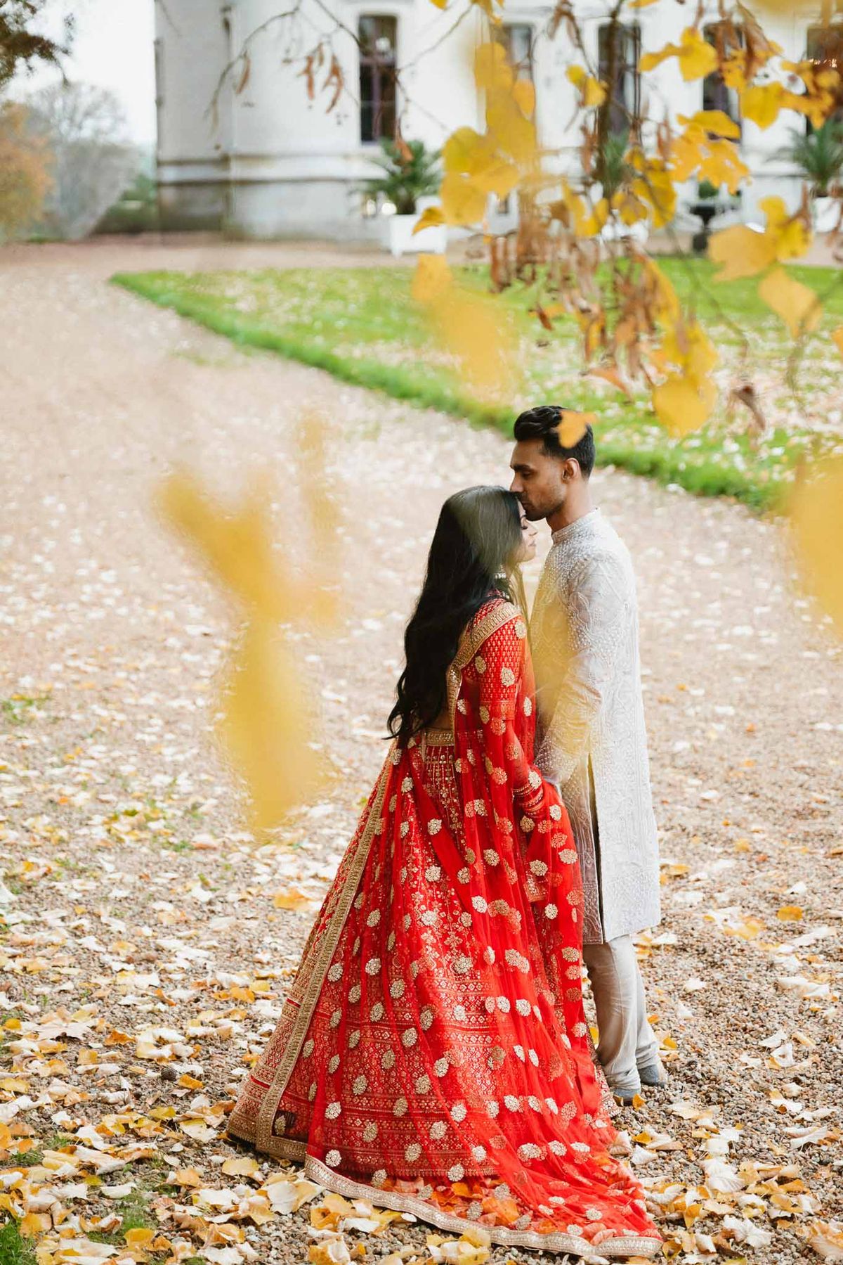 Couple in traditional Indian wedding attire embracing on gravel pathway at European estate in autumn