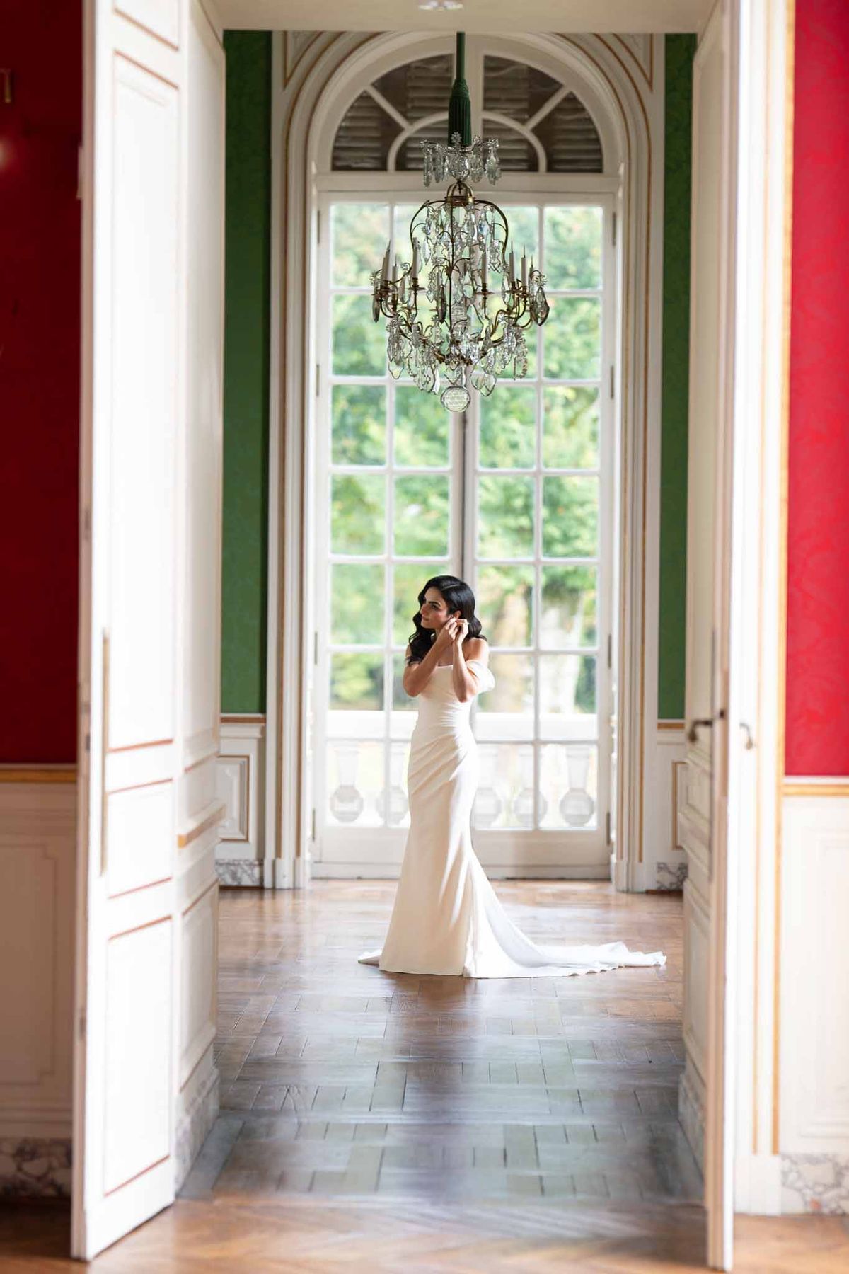 Bride in ivory crepe gown standing in classical gallery hallway with arched windows and chandelier