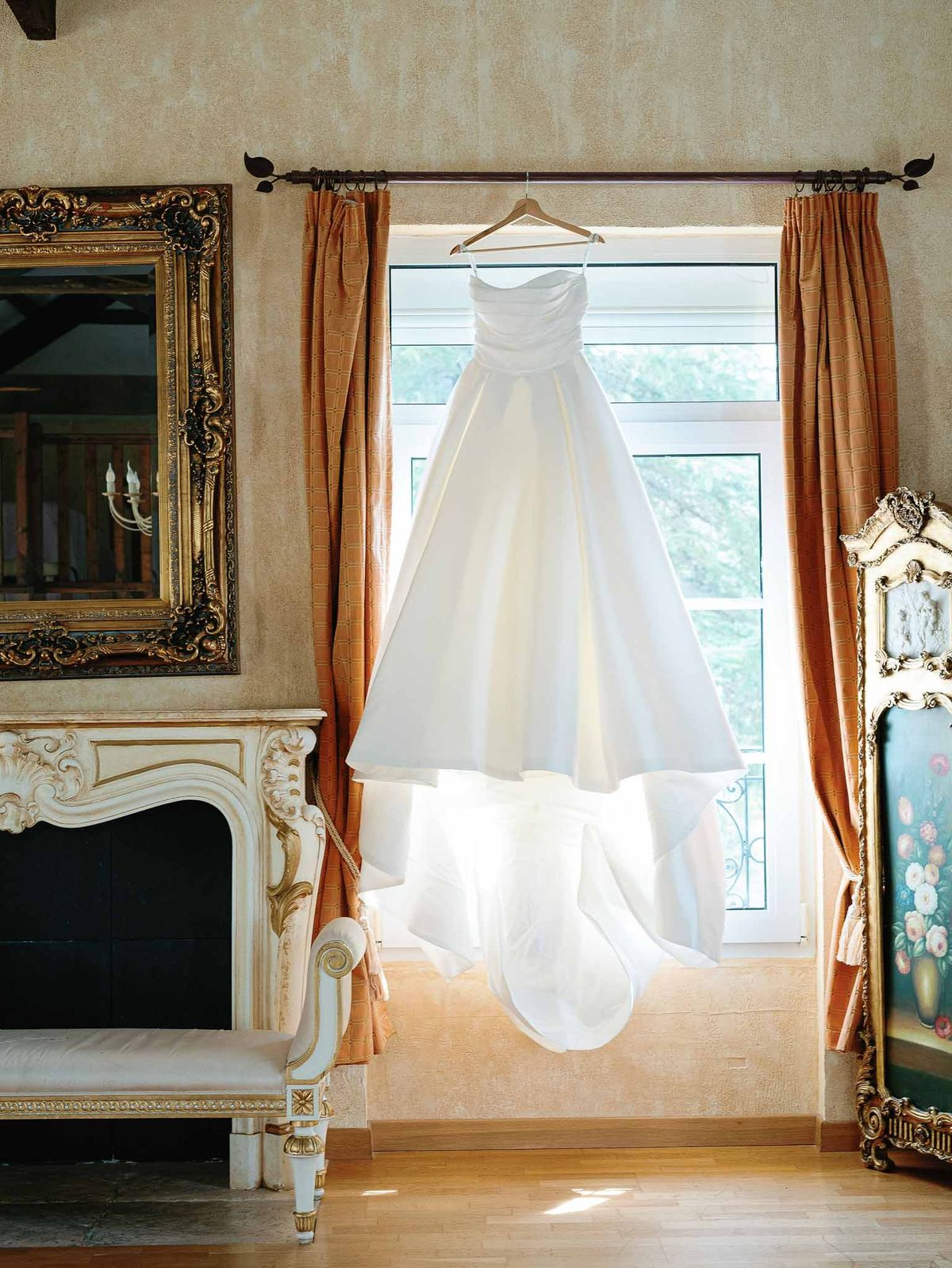 Ivory wedding dress hanging in classic bridal suite with fireplace and gold-framed mirror