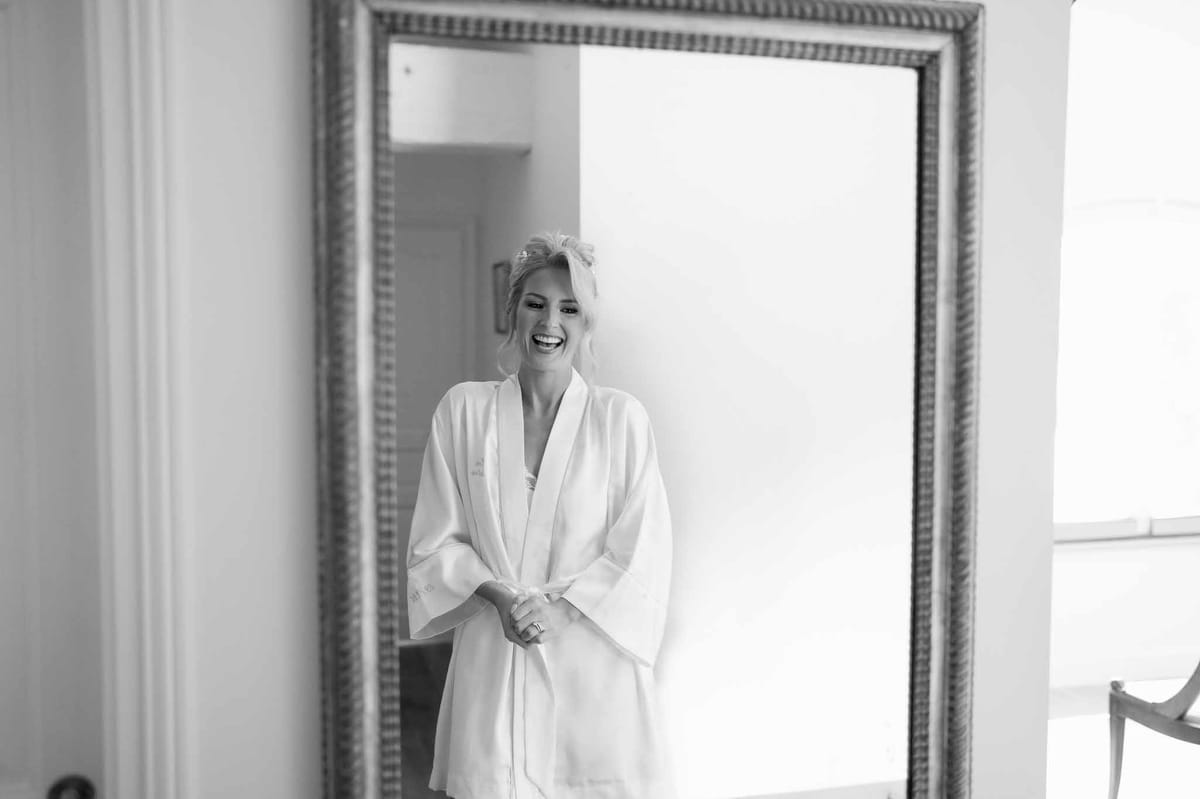 Bride in silk robe smiling during getting ready preparations in bridal suite mirror