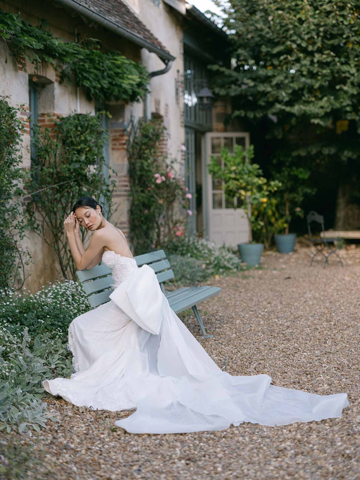 Bride seated on sage bench in courtyard wearing strapless gown with oversized bow and cathedral train