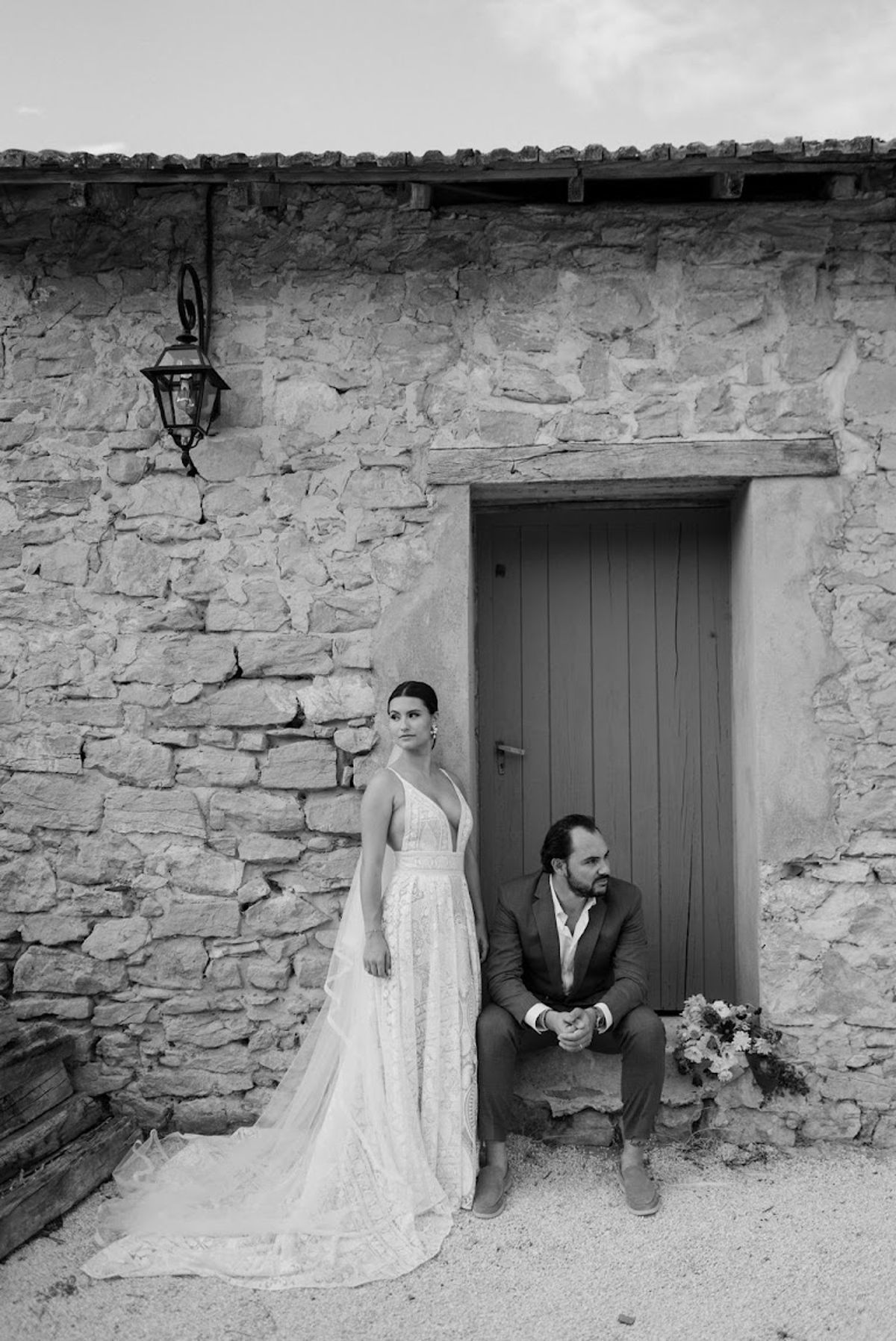 Black and white couple at rustic stone doorway bride in lace gown groom seated on step with bouquet