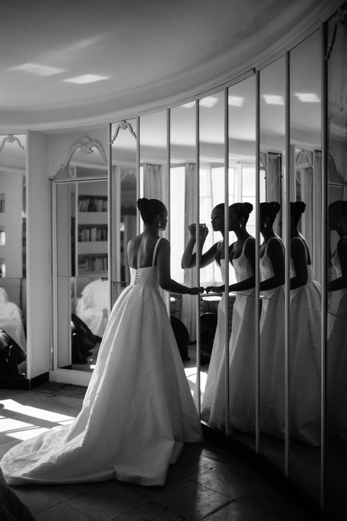 Bride adjusts jewelry before multi-panel mirror with train pooling on floor in ornate white salon in B&W