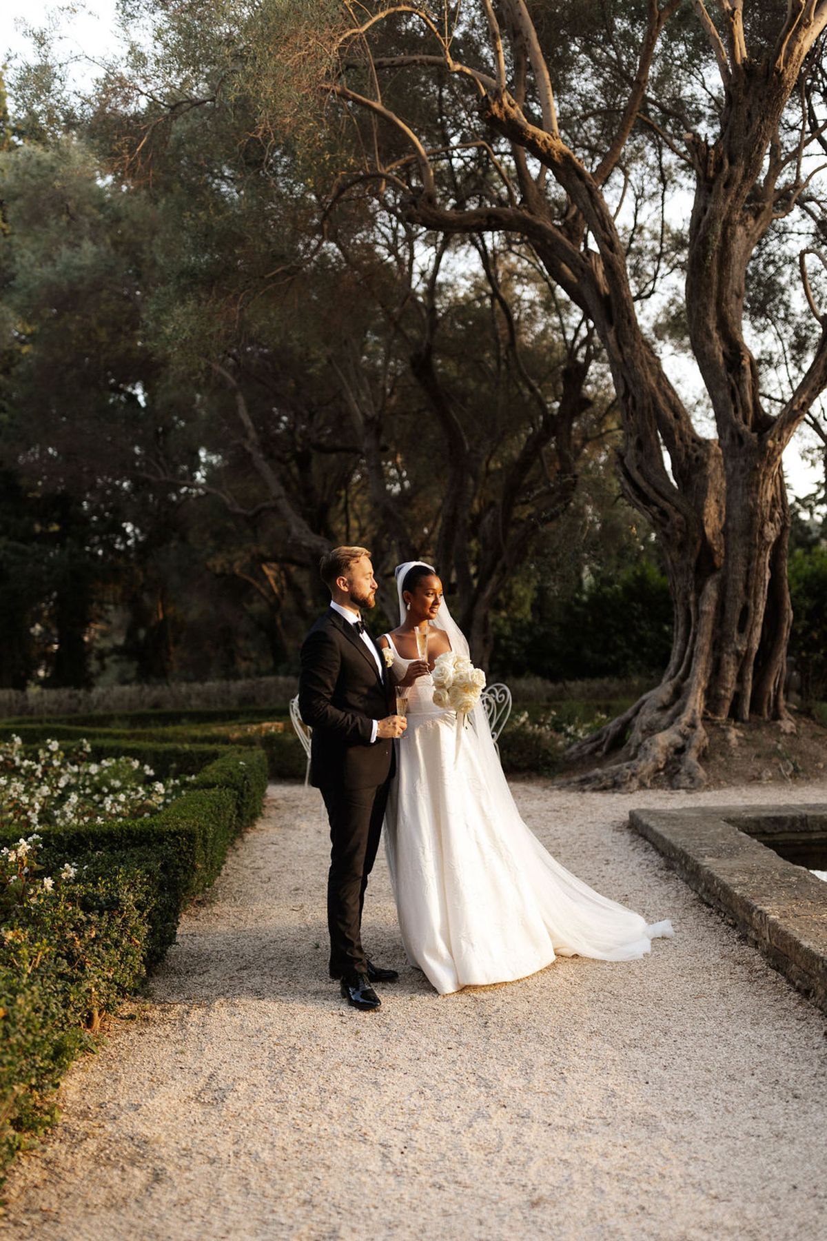 Bride in off-shoulder ivory ballgown and groom in black tuxedo holding champagne in formal garden at golden hour