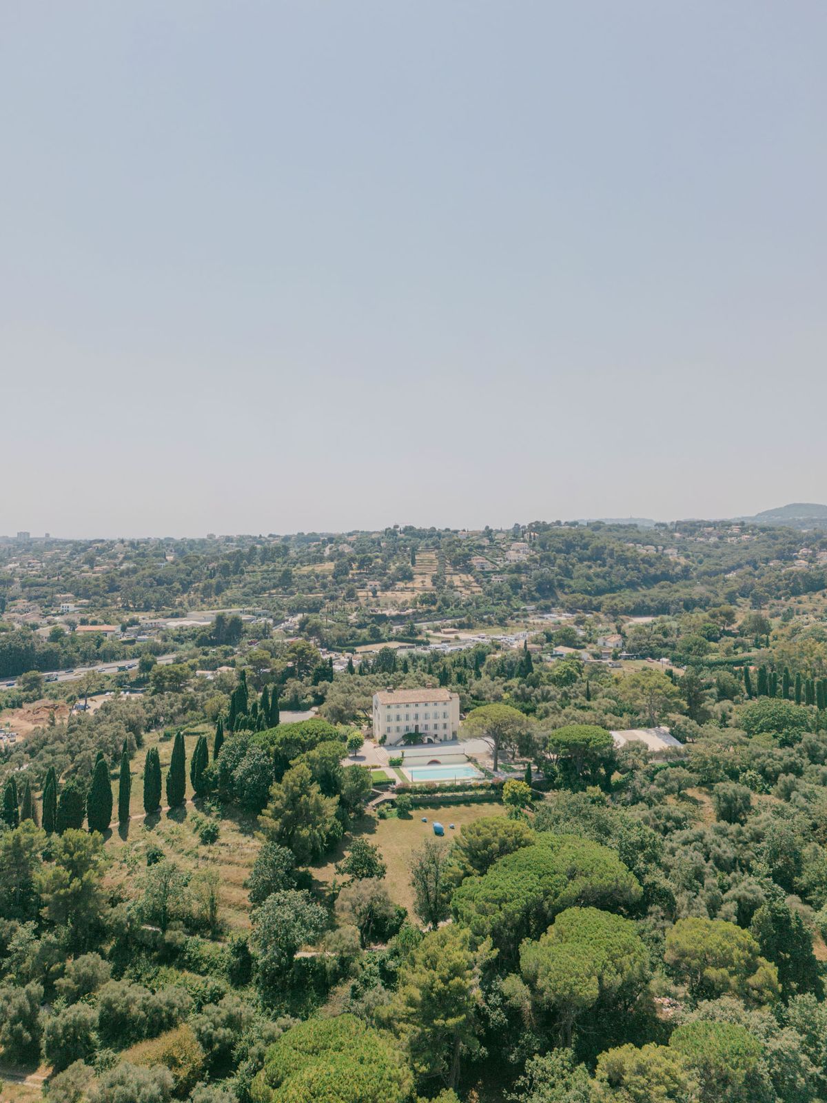 Aerial view of cream chateau with turquoise pool, cypress-lined path, and wooded hillside