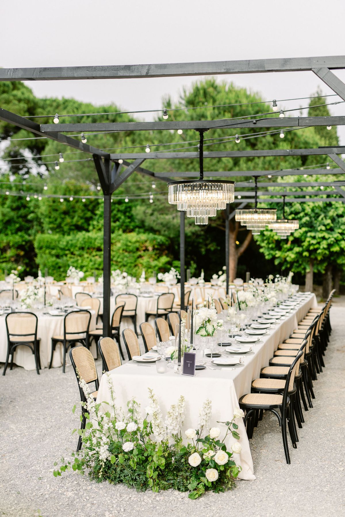 Pergola reception with globe lights, crystal chandeliers, white rose floor arrangements, and cane-back chairs