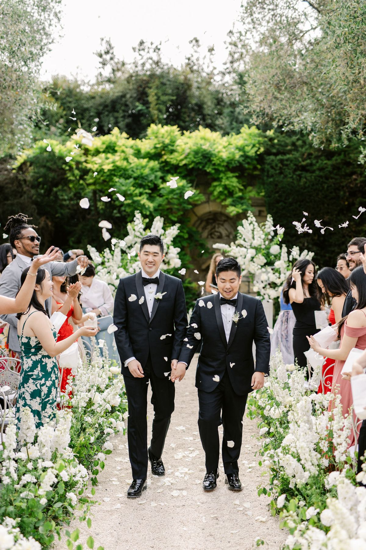 Two grooms in black tuxedos walk aisle as guests toss white petals beside delphinium arrangements