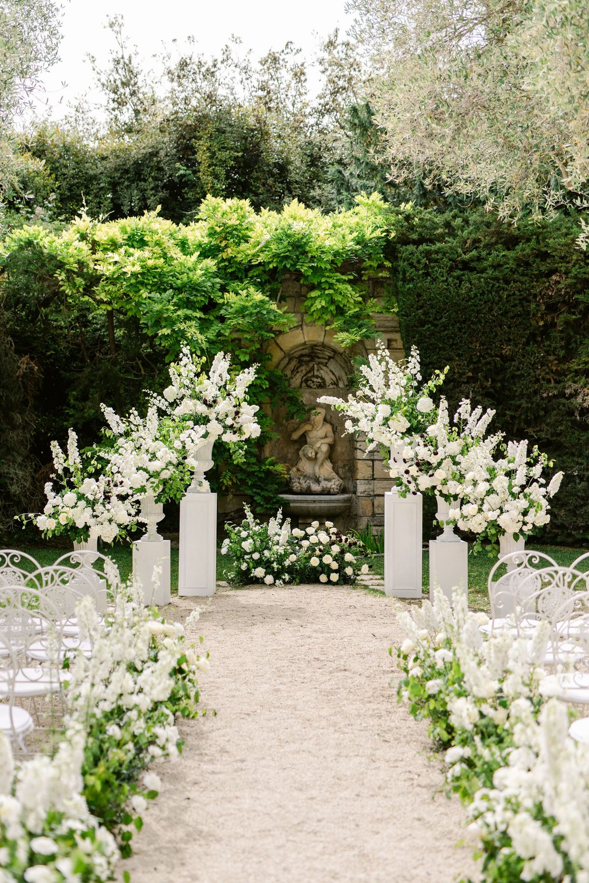 Ceremony setup with white rose aisle arrangements and four pedestal urns before stone fountain niche