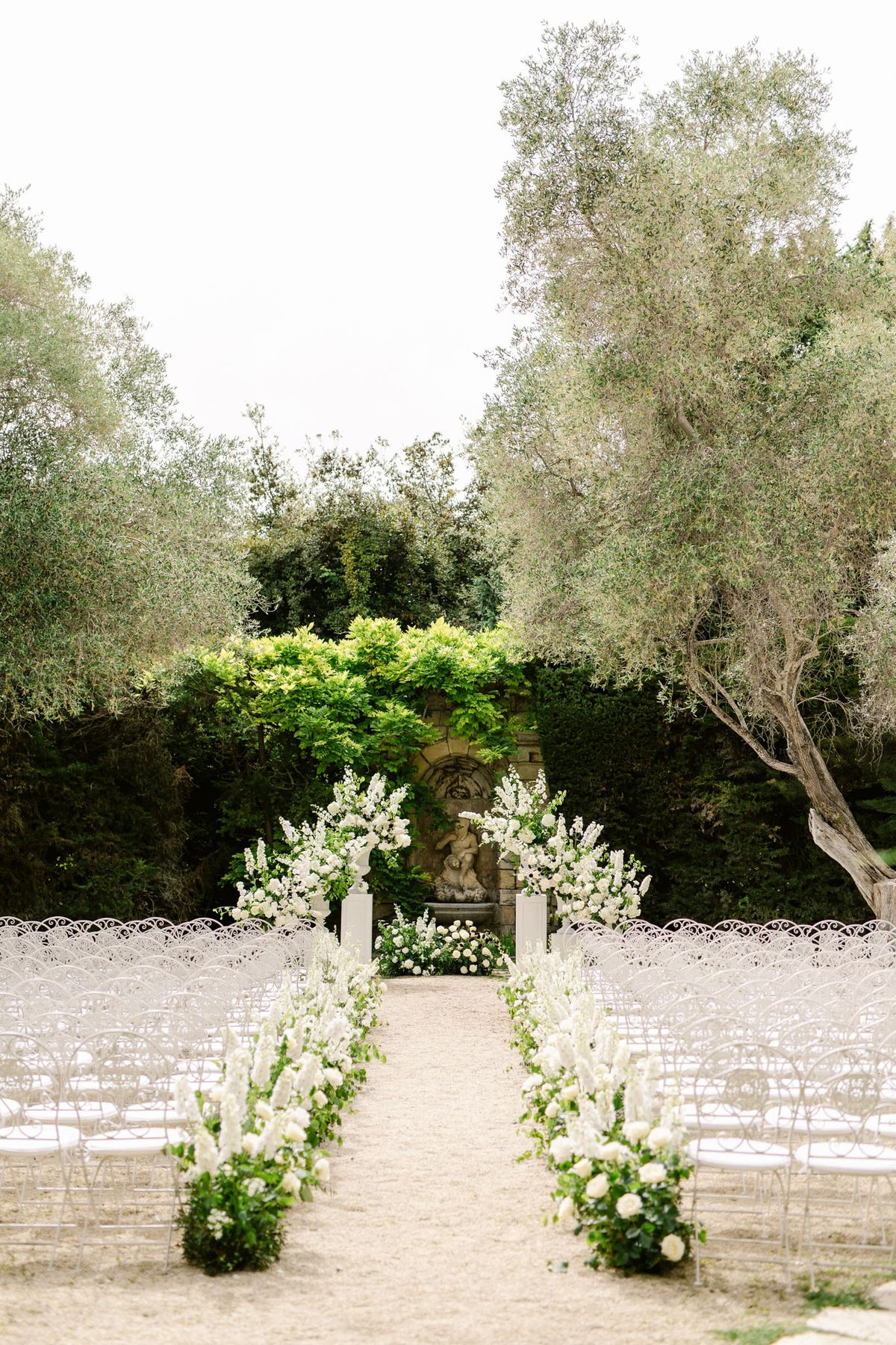 White iron chairs and delphinium aisle columns leading to floral-framed stone fountain altar on lawn