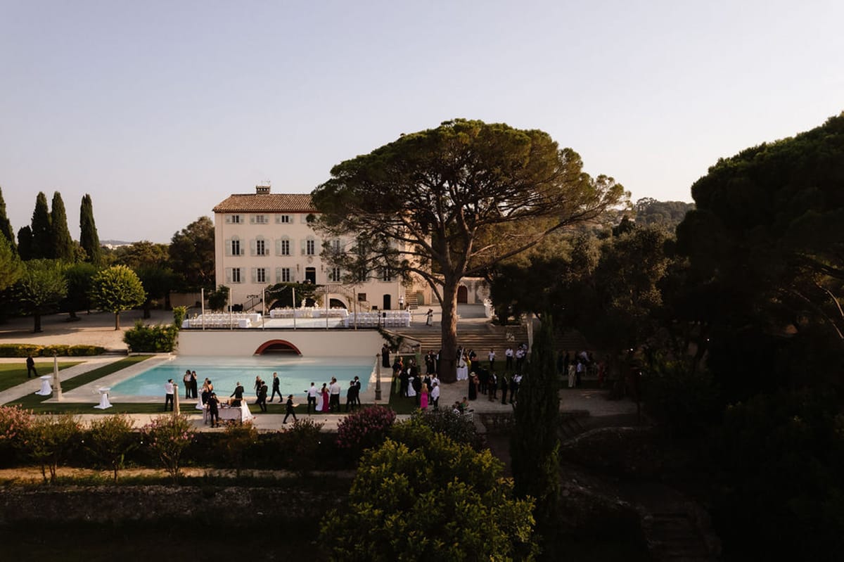 Aerial golden-hour view of 60 guests at poolside cocktail hour before cream bastide with blue shutters