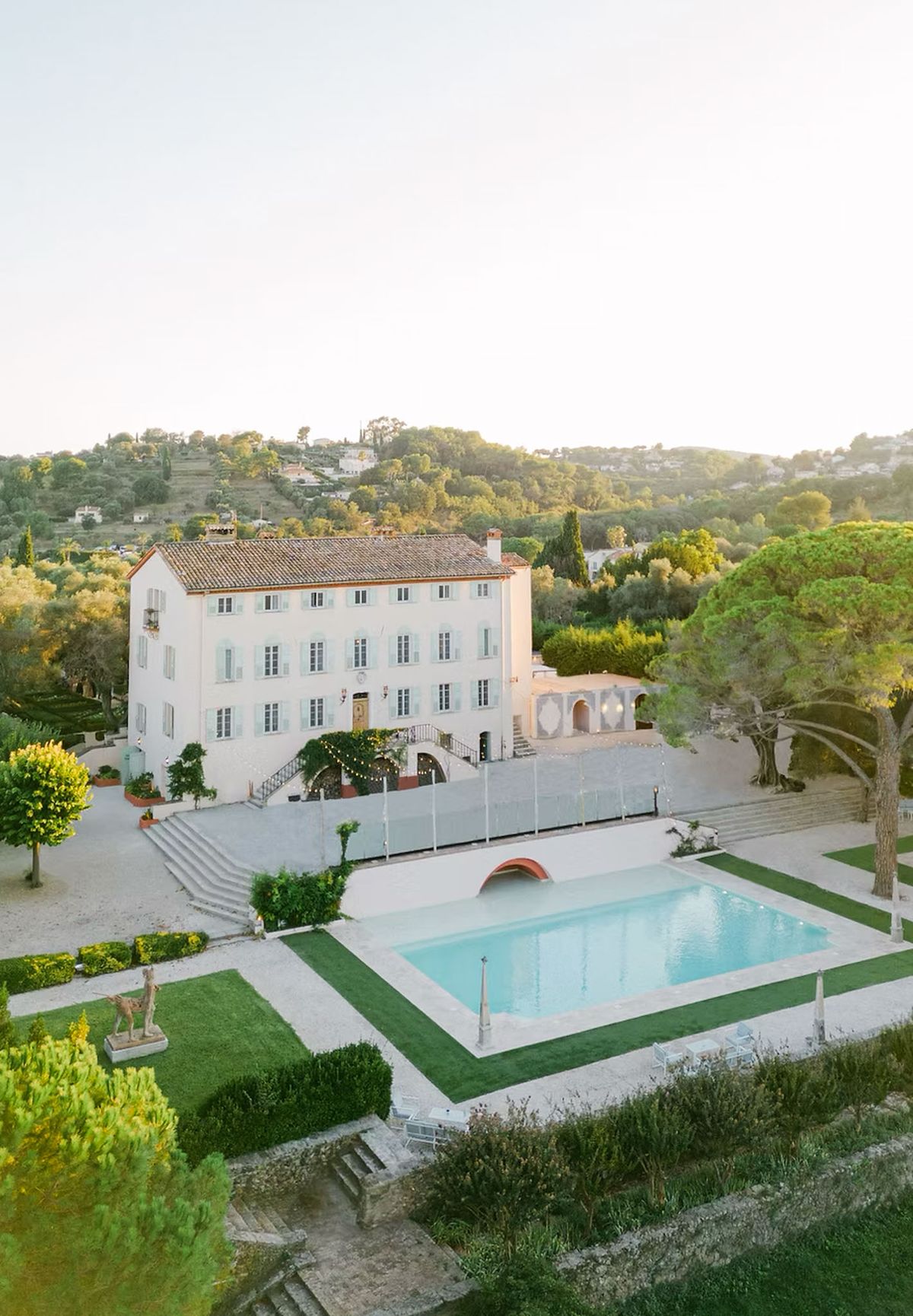 Aerial view of white bastide with pool bridge, parterre gardens, and transparent marquee on hillside