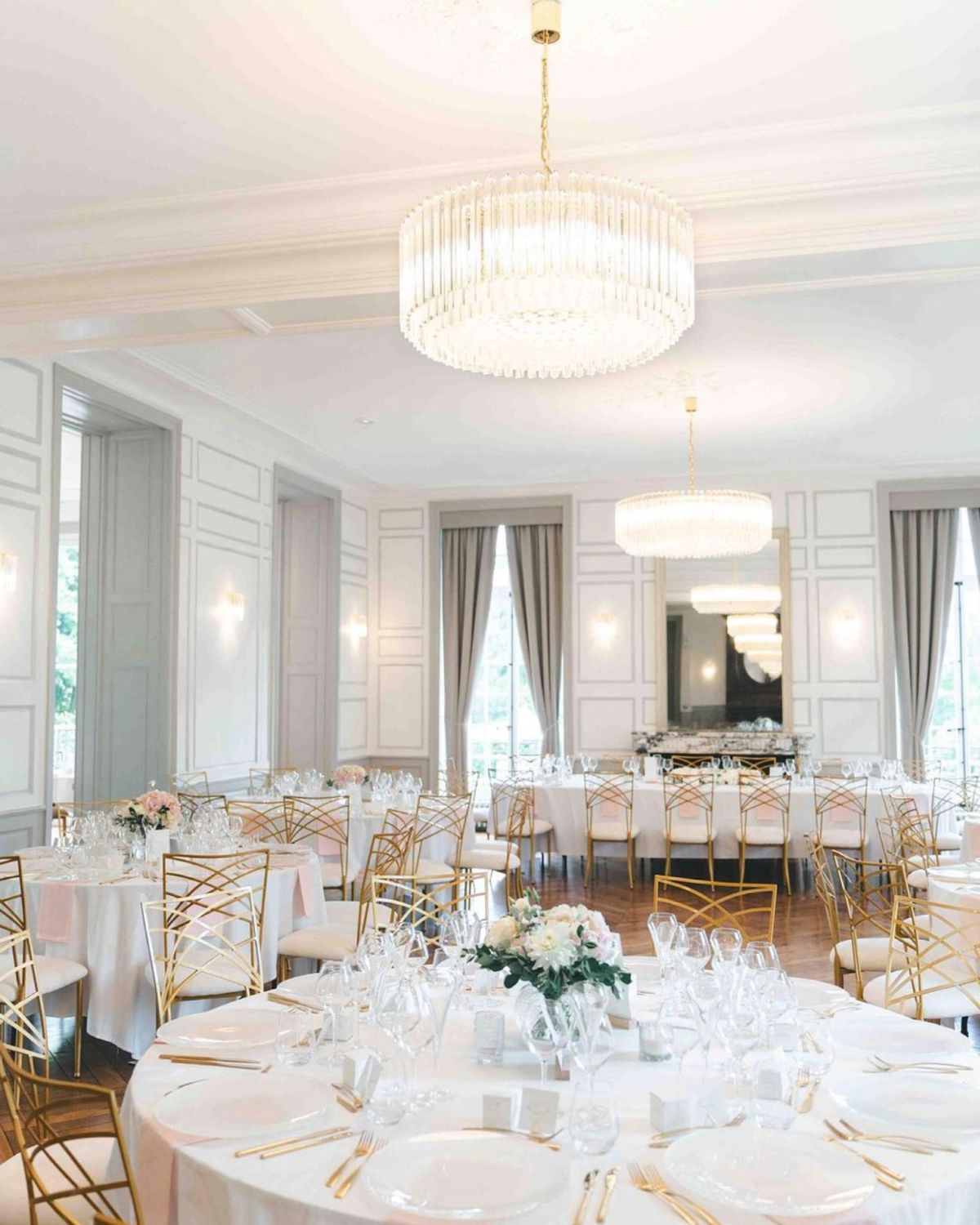 Indoor wedding reception ballroom with round tables, white linens, blush napkins, gold chairs, and crystal chandeliers