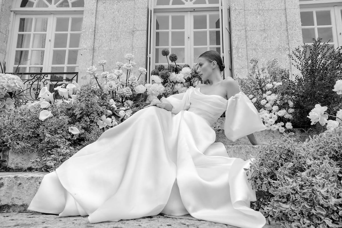 Bride in structured white gown seated among floral arrangements in French courtyard setting