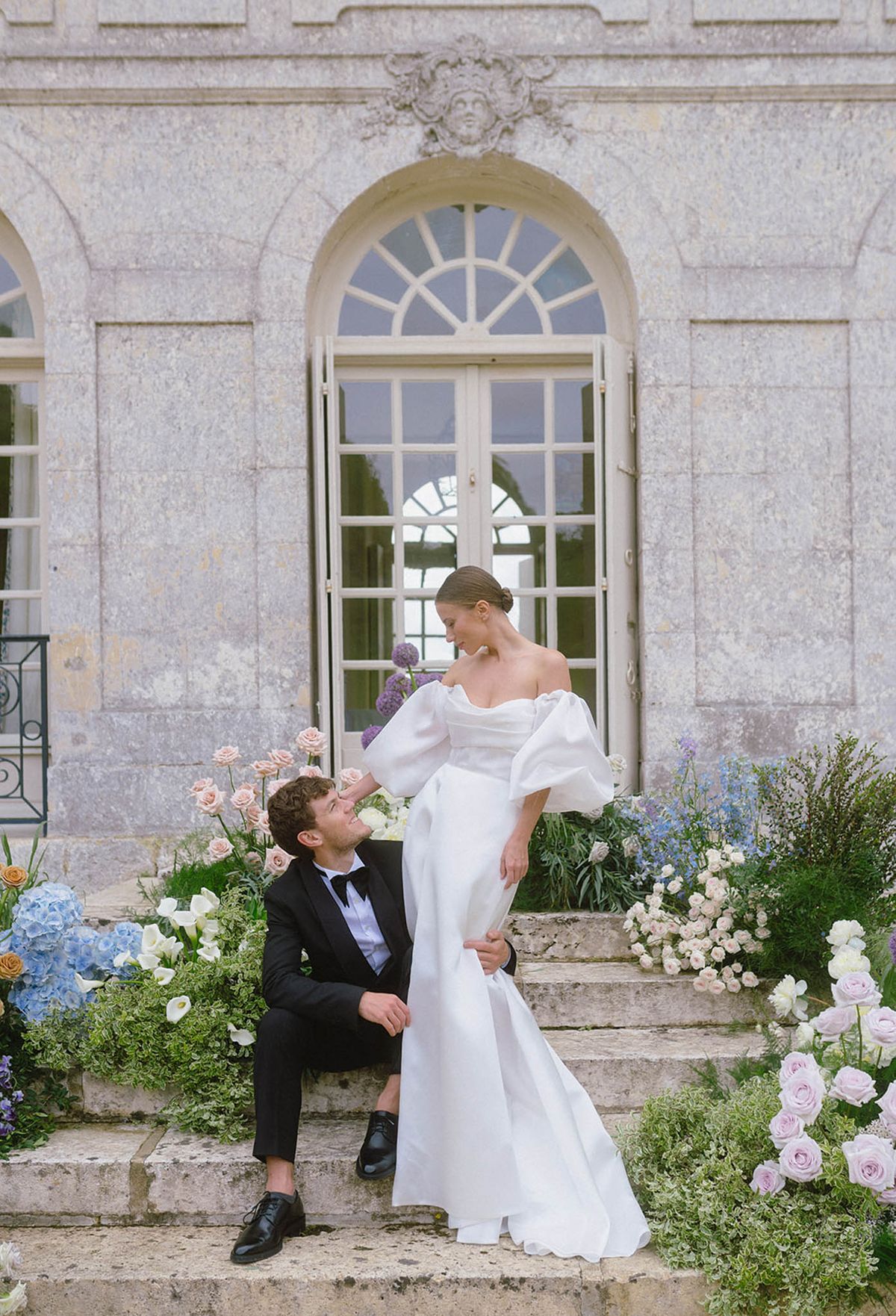 Bride and groom posing on stone steps of classical château with floral arrangements