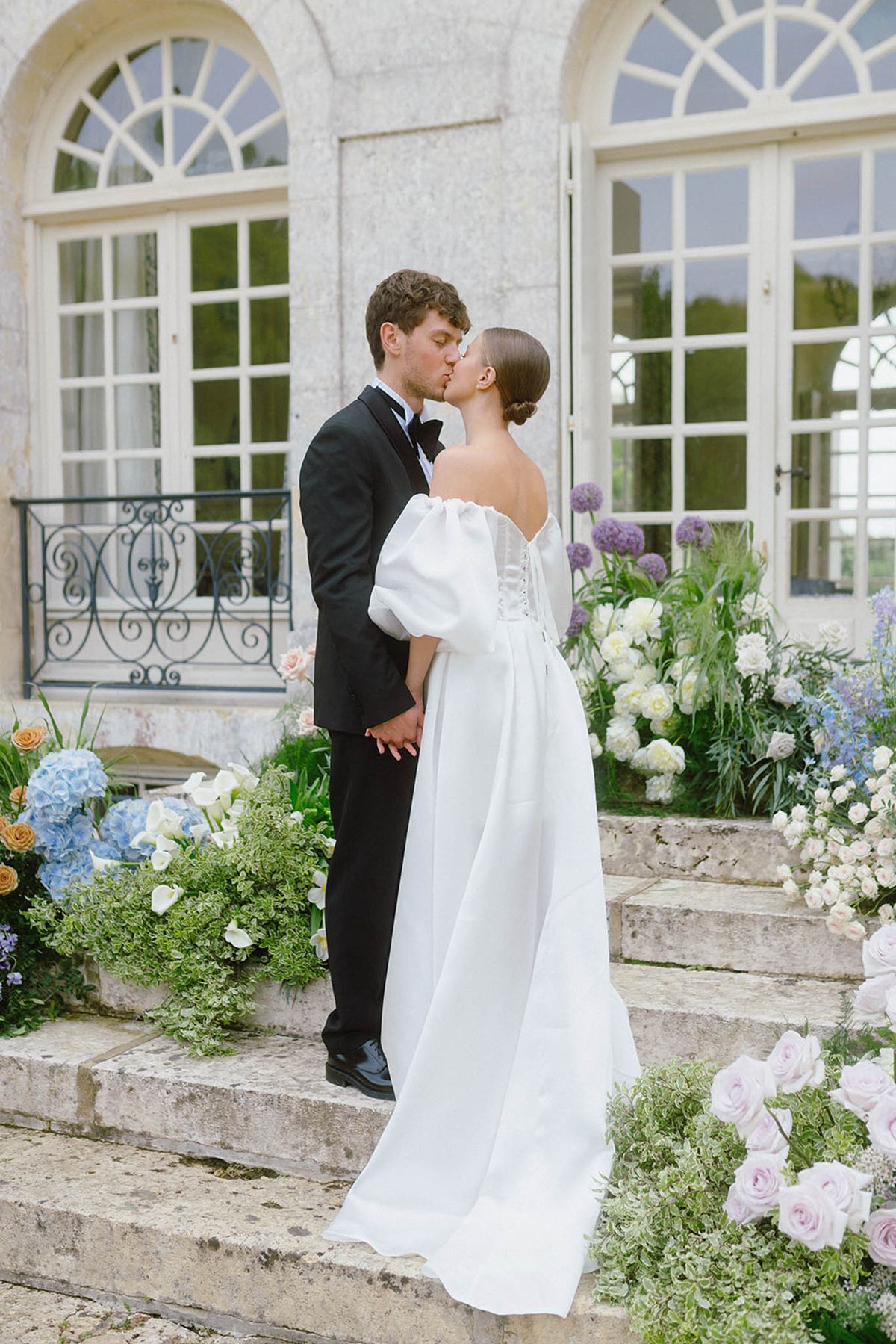 Bride and groom kissing on stone terrace of classical manor house with floral arrangements